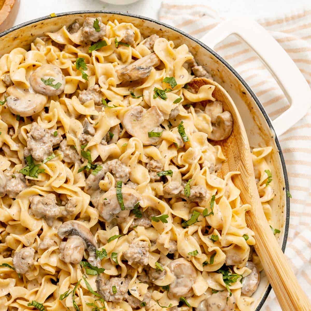 A skillet filled with creamy egg noodles, ground meat, mushrooms, and garnished with chopped parsley, with a wooden spoon resting on the side.