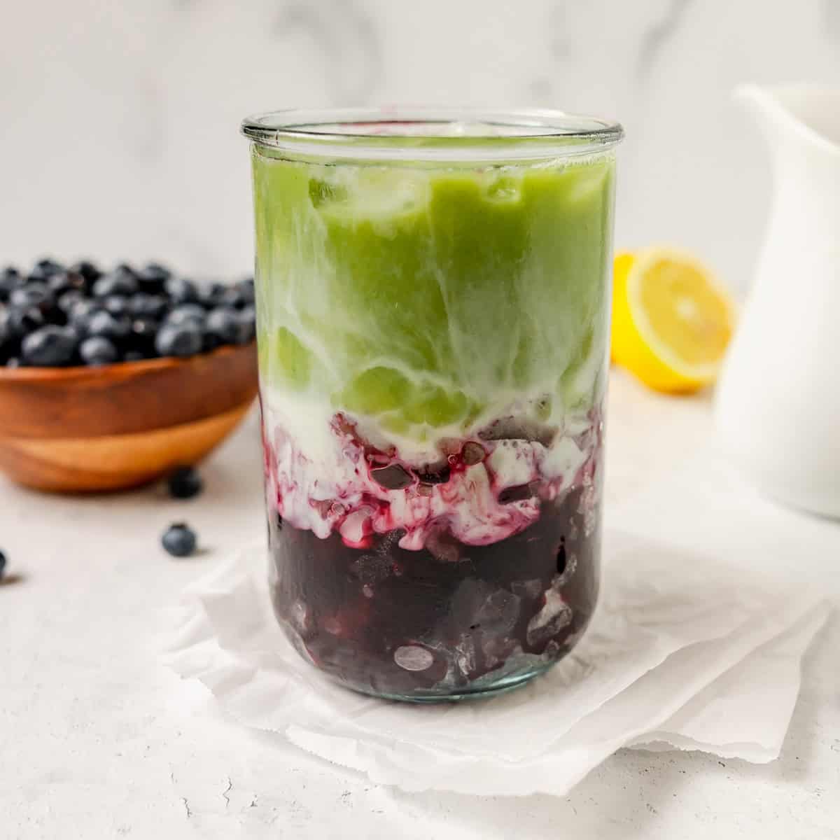 A glass of iced matcha latte with layers of milk and blueberry puree, placed on a napkin with fresh blueberries and a lemon in the background.