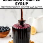 A glass bottle of Blueberry Coffee Syrup with a pour spout sits on a woven mat. A bowl of blueberries and a white pitcher are in the background.