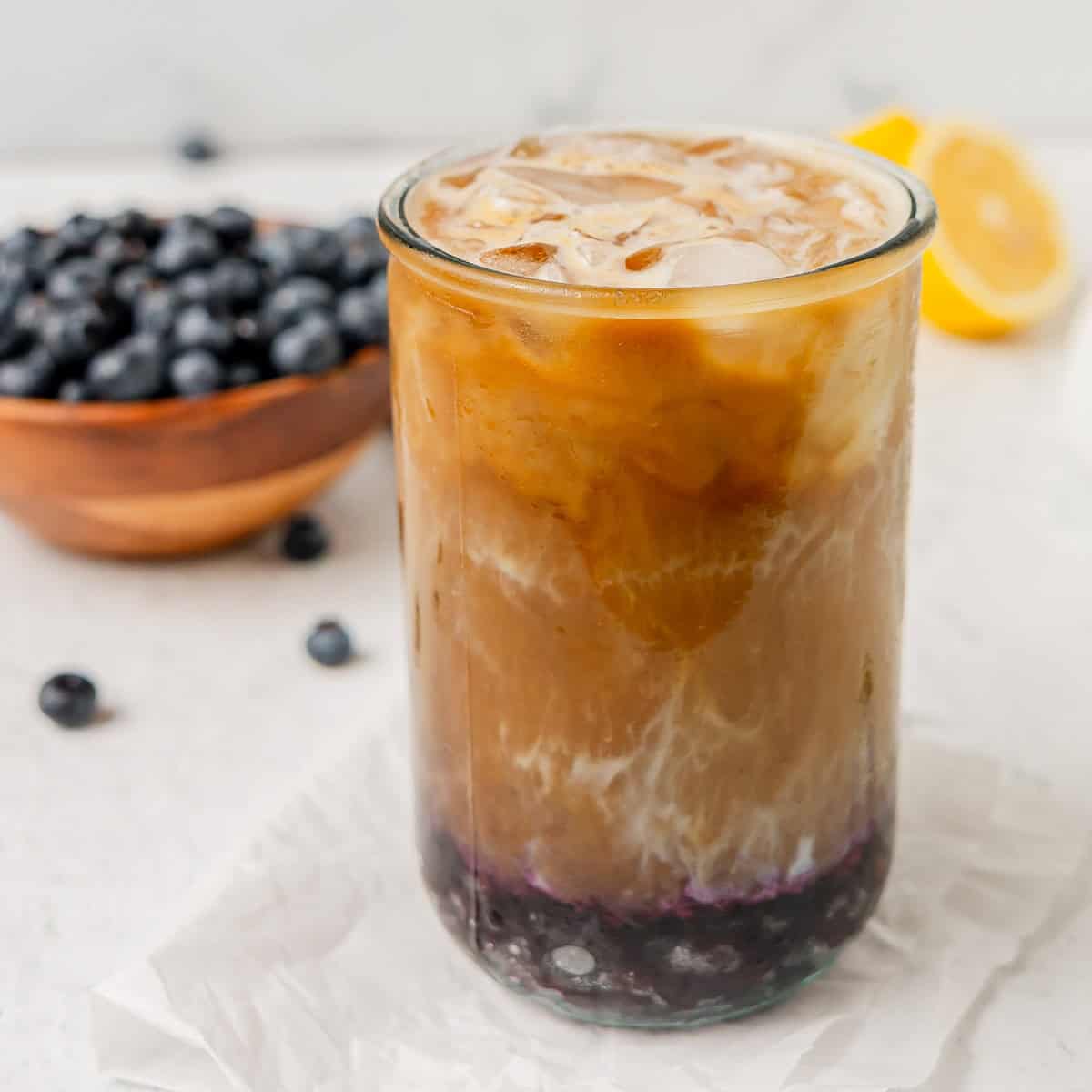 A glass of iced coffee with milk and blueberry syrup at the bottom, with ice cubes on top. A bowl of blueberries and a lemon are in the background.