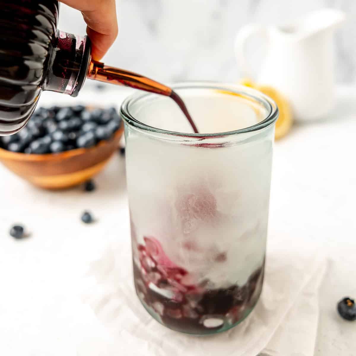 Hand pouring blueberry syrup into a glass of ice, with a bowl of blueberries and a white pitcher in the background.