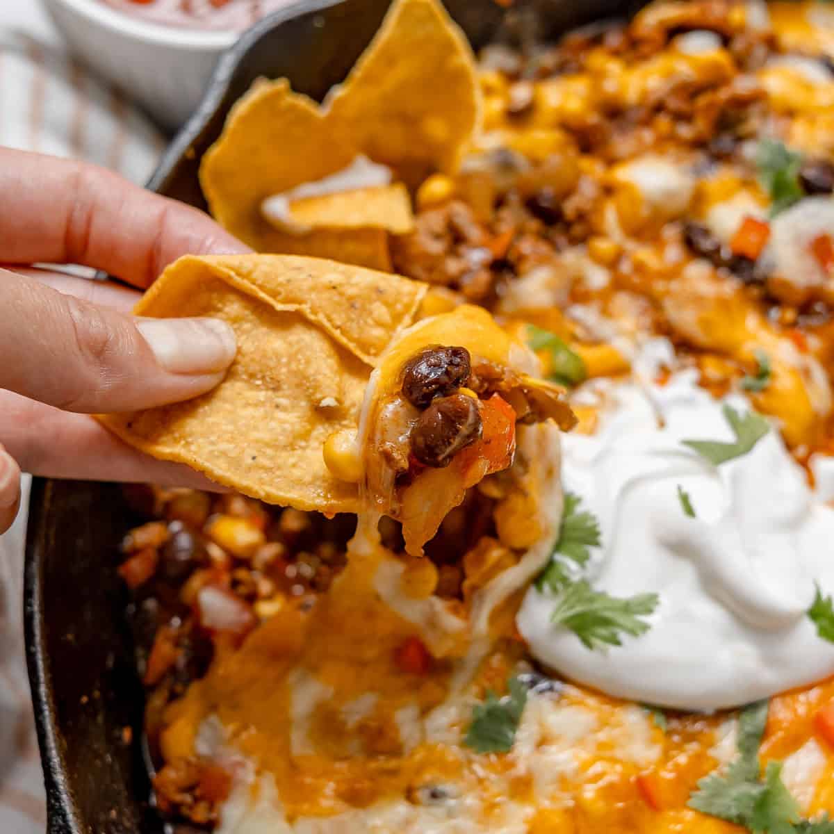 A hand dips a tortilla chip into a skillet of cheesy black bean and corn dip topped with sour cream and cilantro.