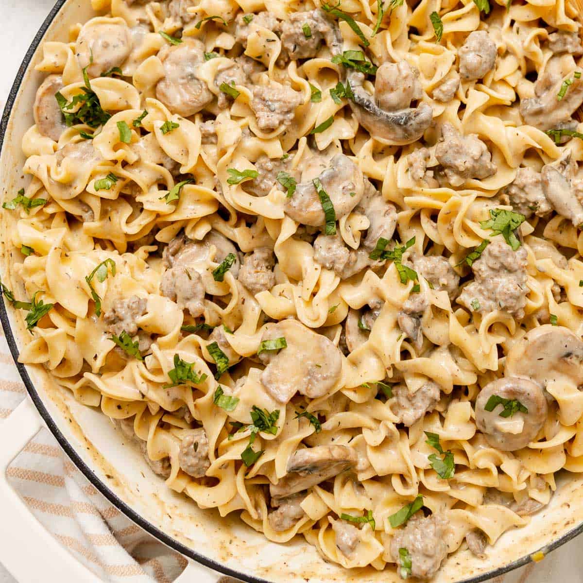 A skillet filled with egg noodles, ground meat, sliced mushrooms, and a creamy sauce, garnished with chopped parsley.
