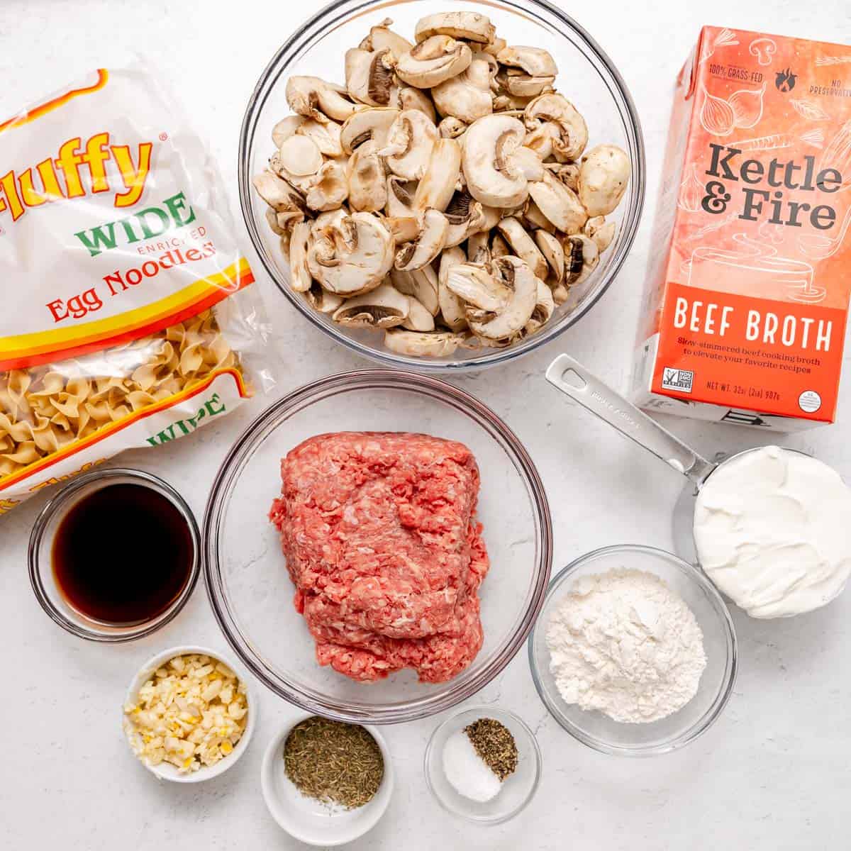 Ingredients for beef stroganoff arranged on a white surface: ground beef, sliced mushrooms, egg noodles, beef broth, sour cream, chopped garlic, flour, black pepper, dried thyme and soy sauce.