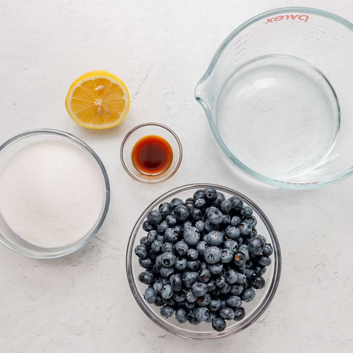 Overhead view of bowls containing blueberries, sugar, lemon half, vanilla extract, and a measuring cup of water, all arranged on a white surface.
