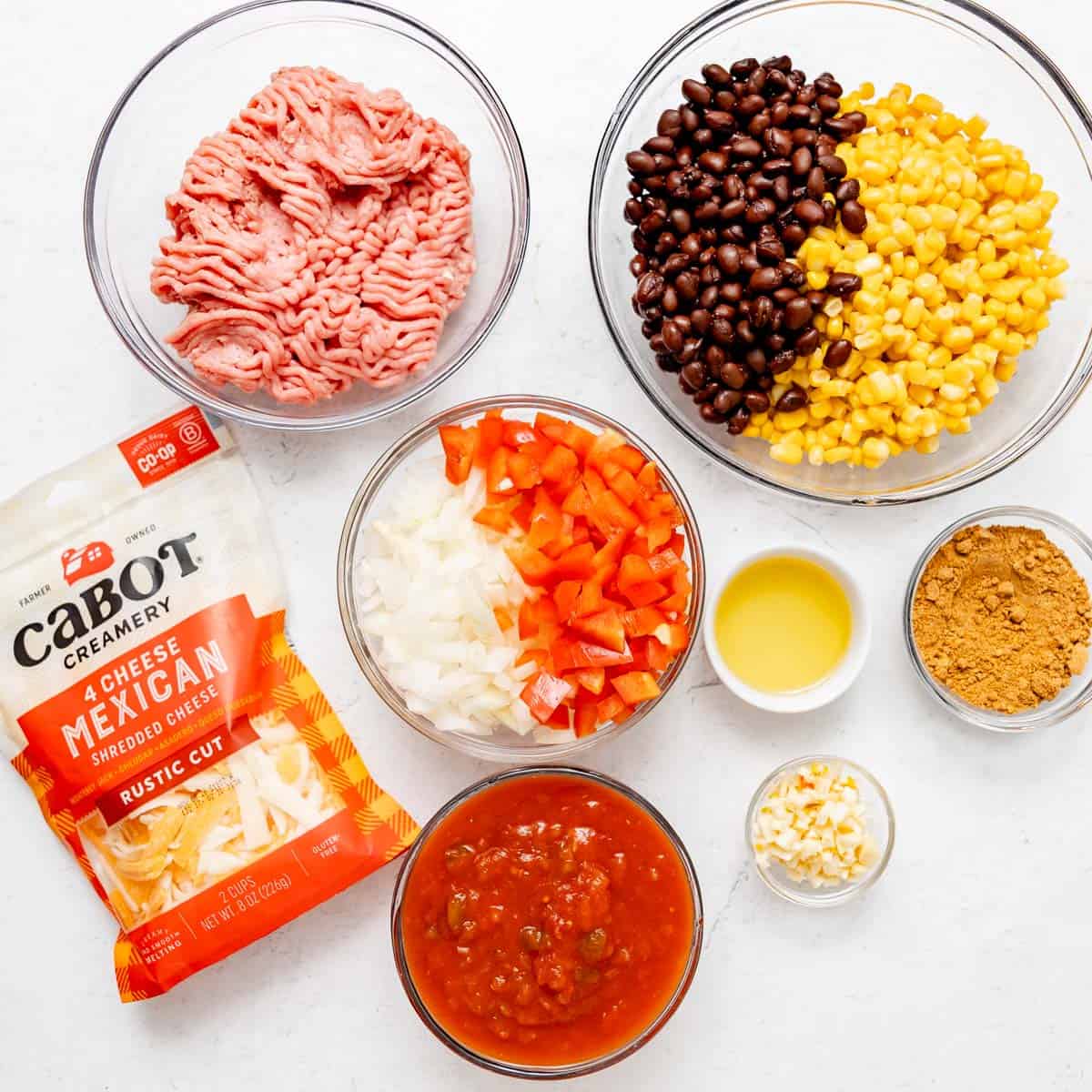 Bowls of ground turkey, black beans with corn, chopped onions and peppers, salsa, oil, taco seasoning, minced garlic, and a bag of shredded Mexican cheese arranged on a white surface.