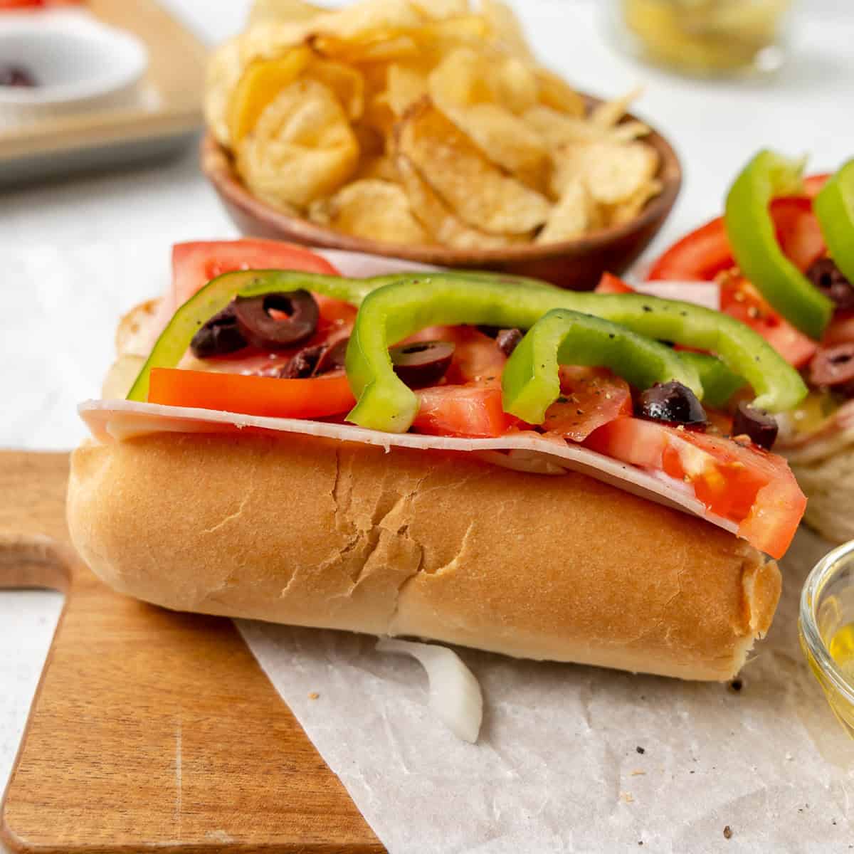 A sub sandwich with ham, tomato, green bell pepper, and purple olives on a wooden board, with potato chips in the background.