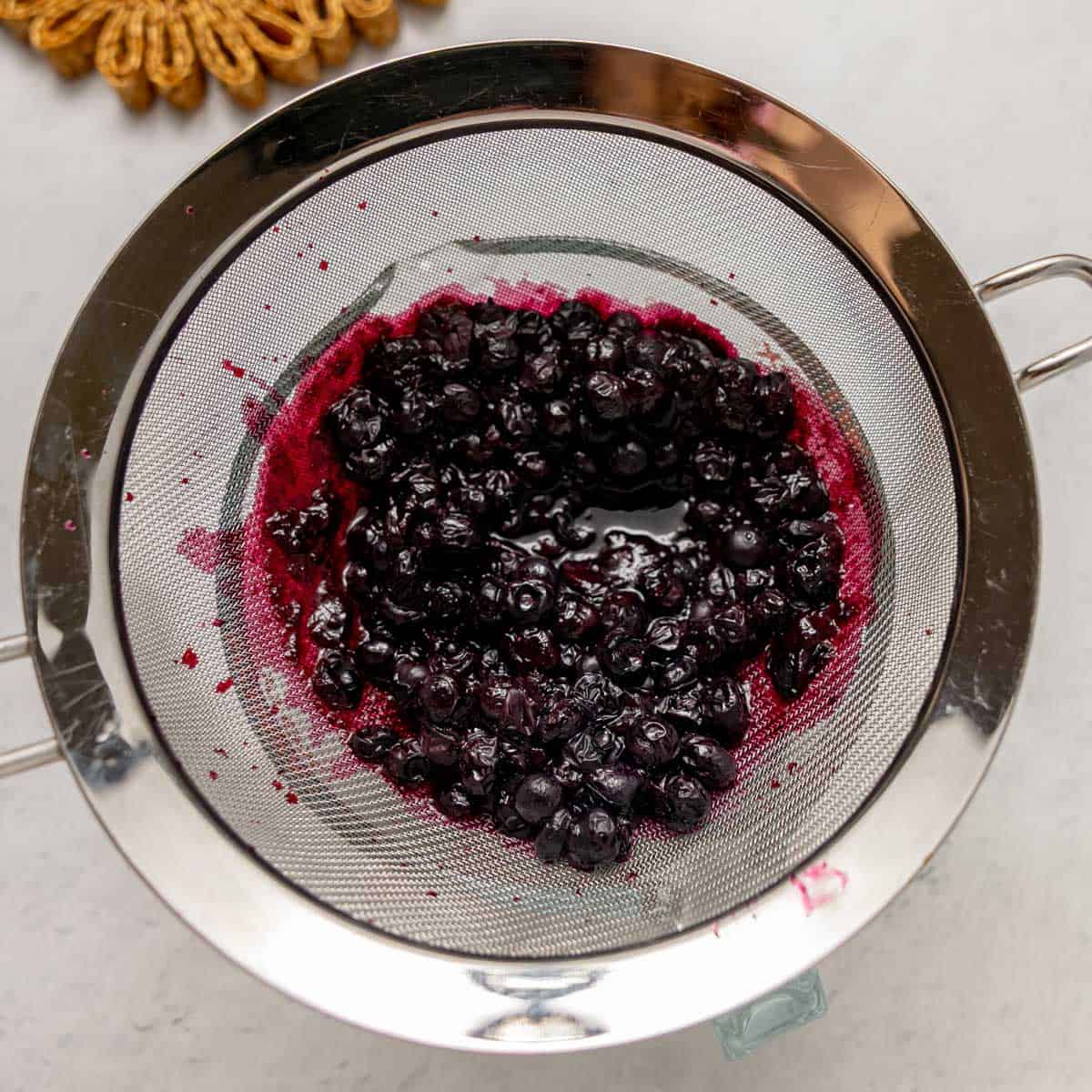 A metal strainer holds cooked blueberries, with juice and pulp visible, placed on a light-colored surface.