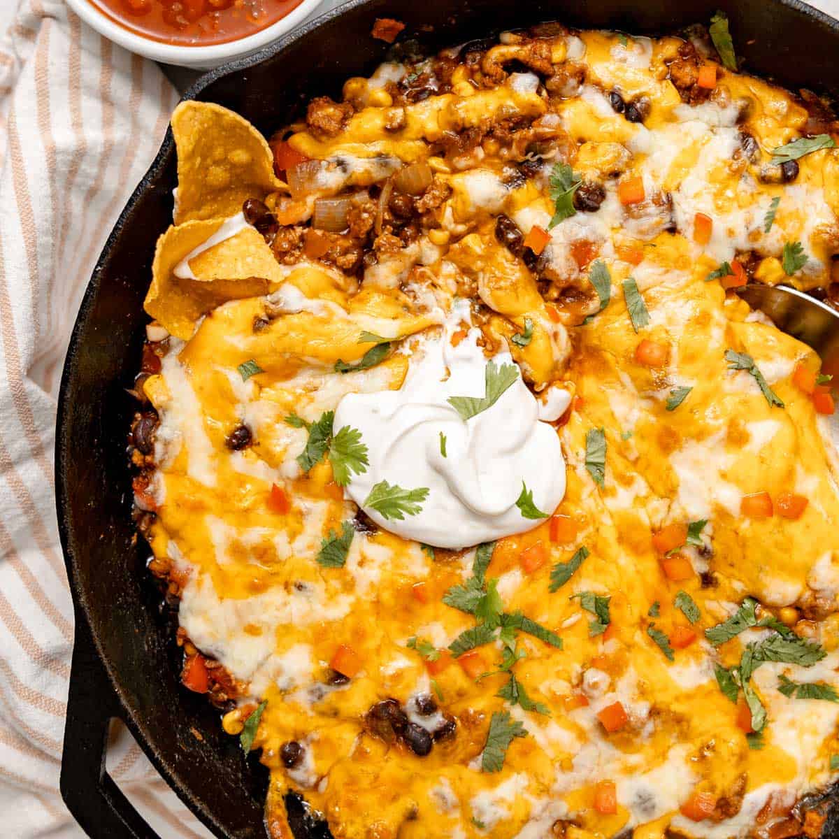 A skillet filled with cheesy taco meat topped with sour cream, chopped tomatoes, cilantro, and tortilla chips, with a small bowl of salsa nearby.