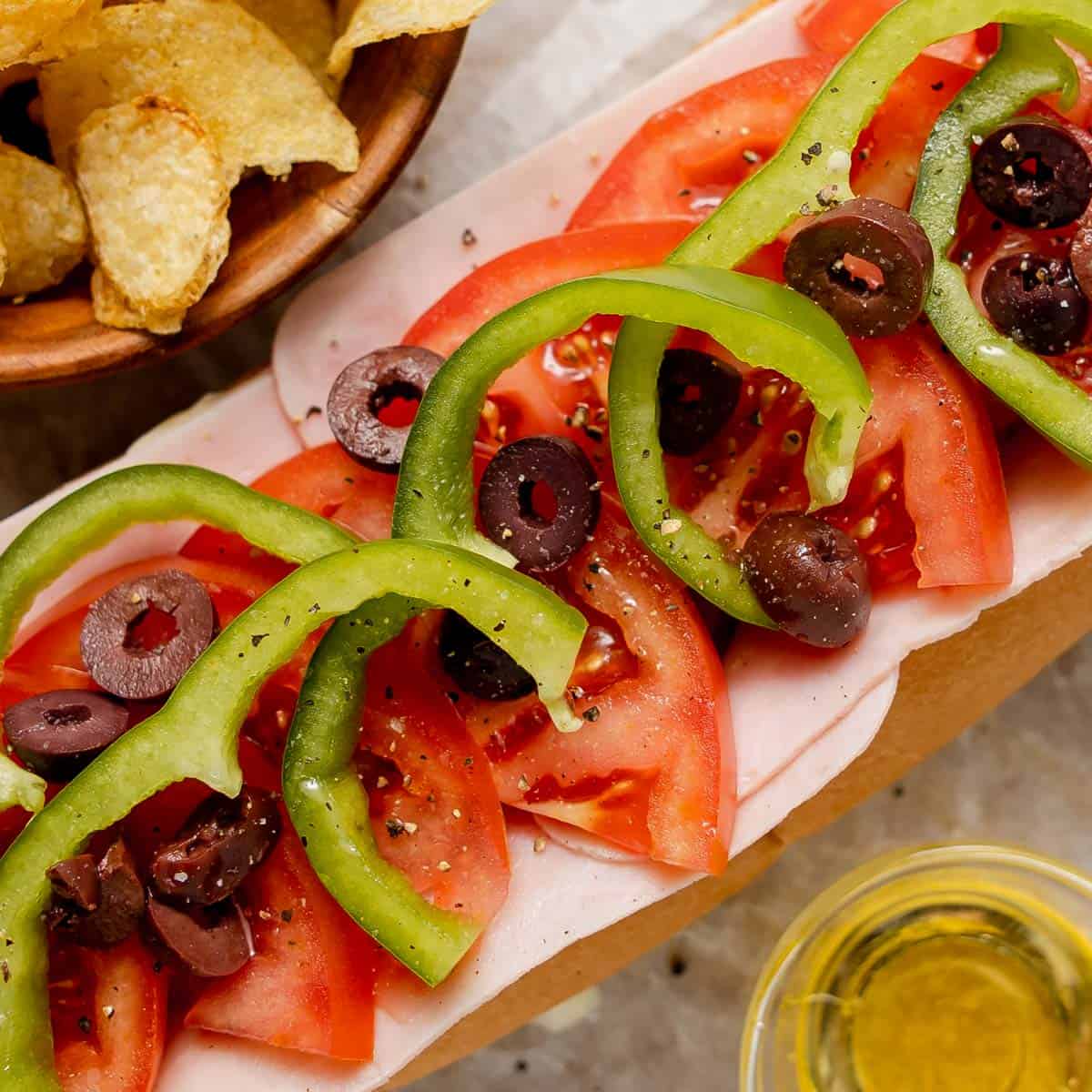 Sub sandwich topped with sliced tomatoes, green bell peppers, black olives, and black pepper, served with a side of potato chips and a small cup of yellow condiment.