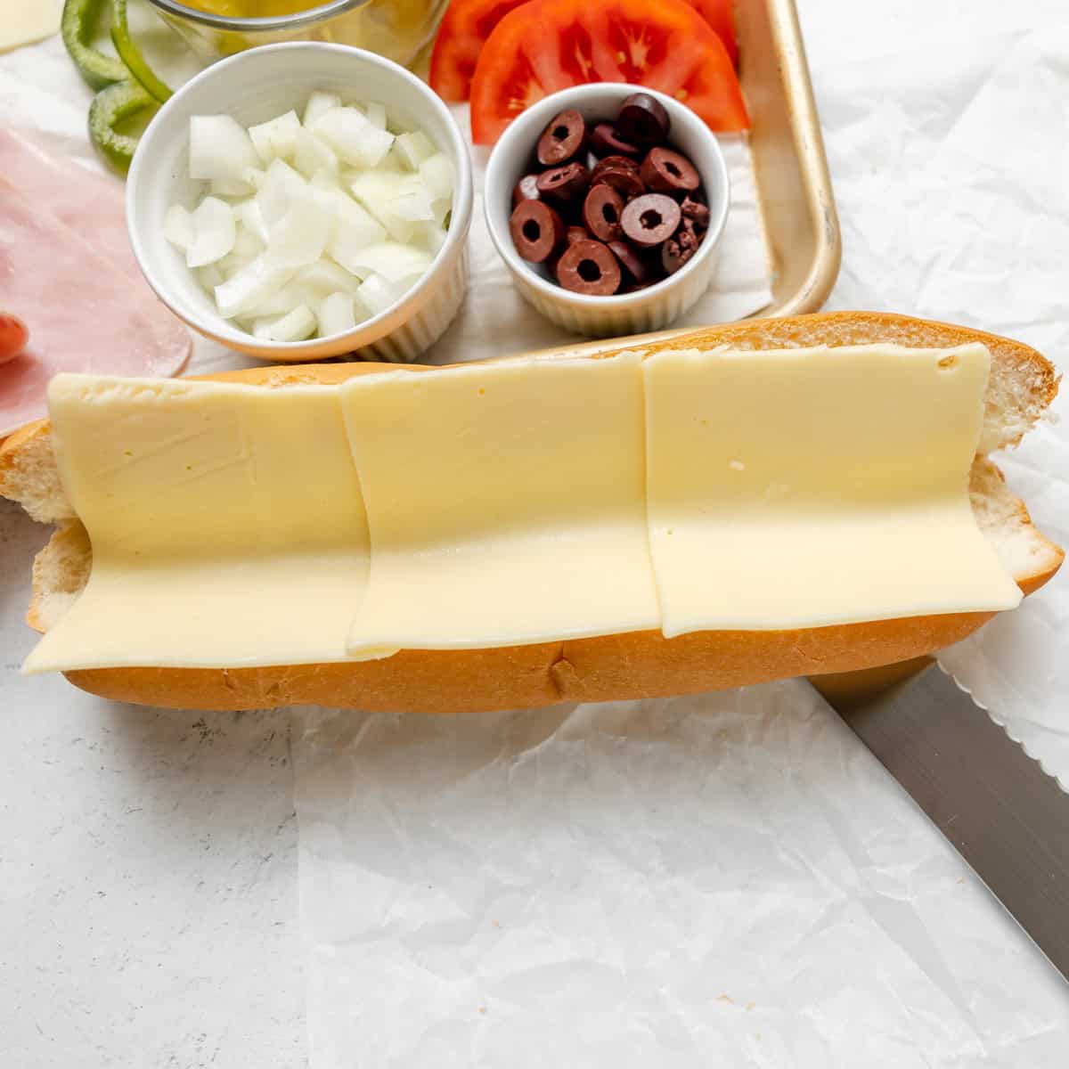 A hoagie roll with three slices of cheese, next to a tray holding chopped onions, sliced black olives, tomato, and other sandwich ingredients.