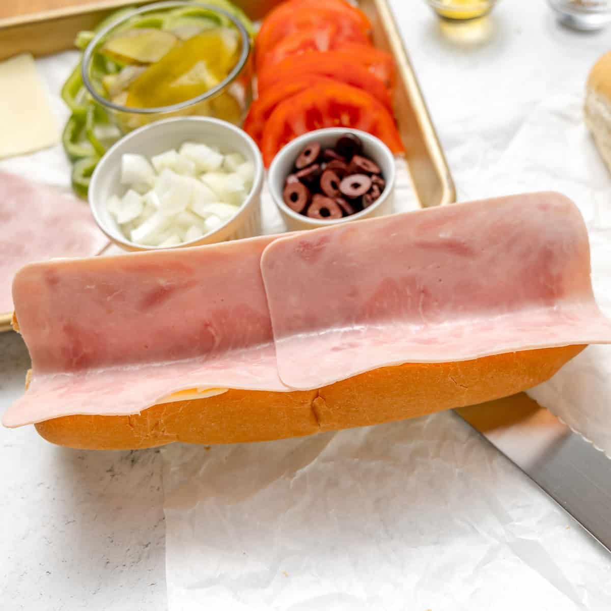 An open sandwich roll with two slices of ham, beside bowls of sliced pickles, tomatoes, onions, and olives on a tray.