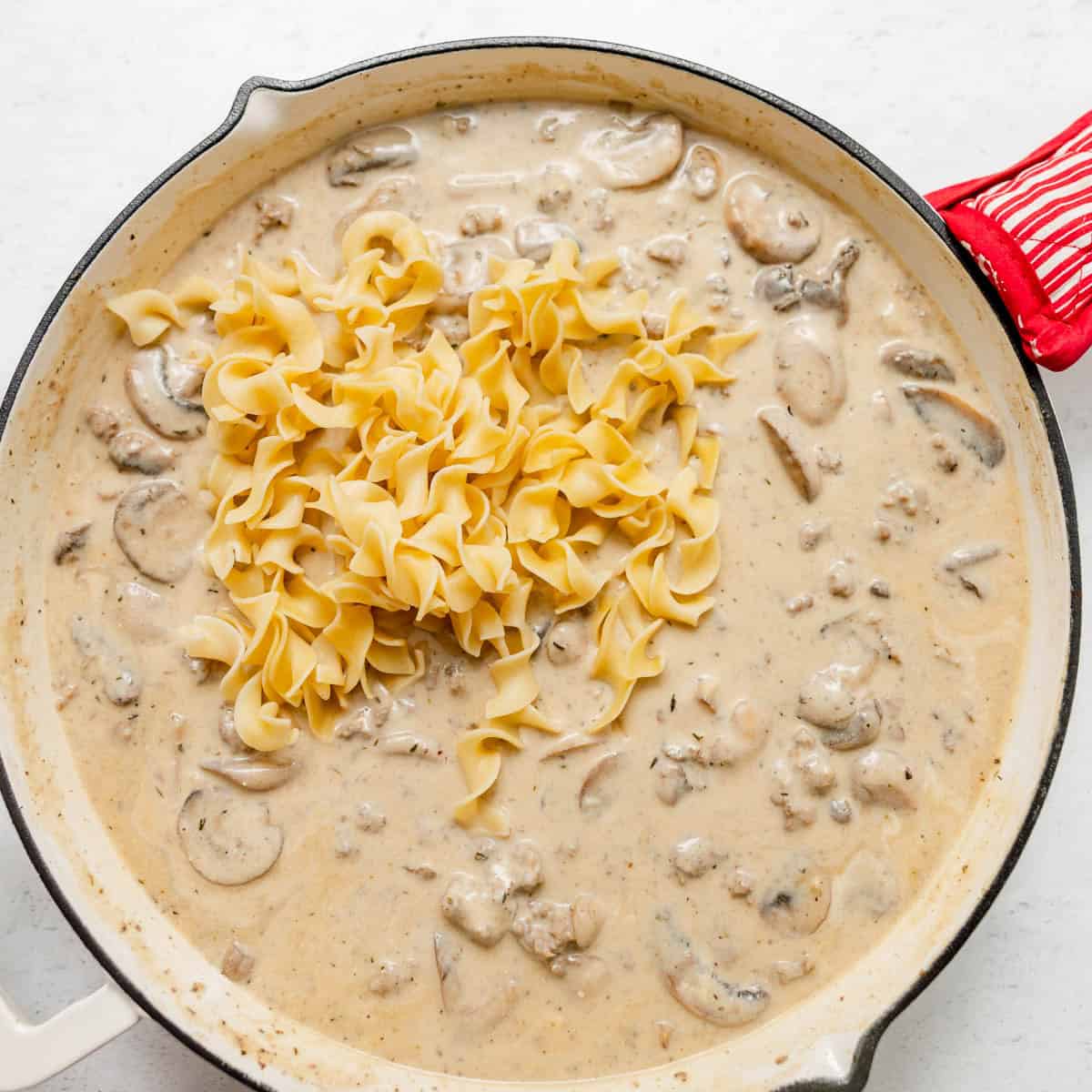 A skillet filled with creamy beef stroganoff sauce with mushrooms and a pile of egg noodles on top, ready to be mixed in.