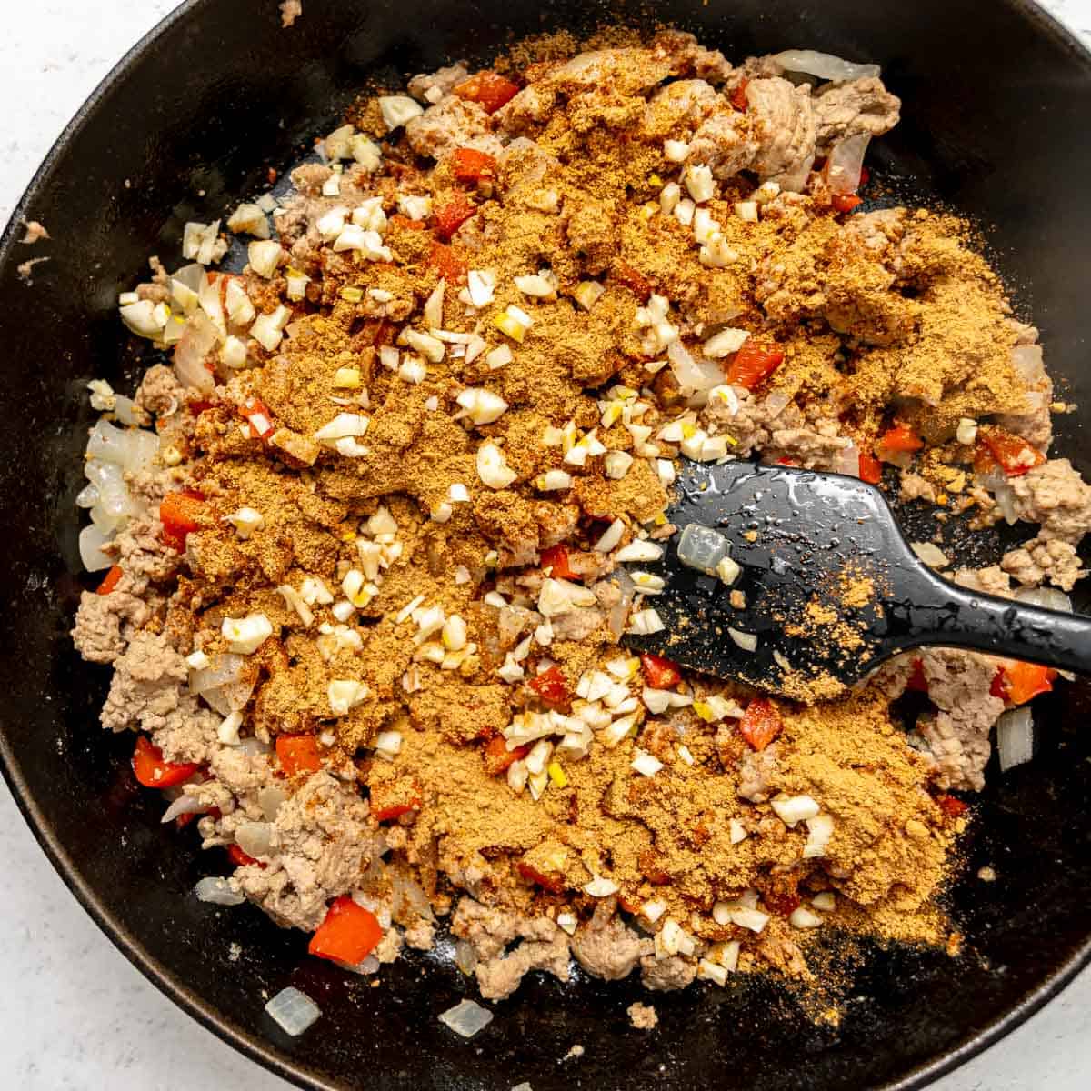 Ground meat, diced onions, red peppers, chopped garlic, and spices are being cooked and mixed in a black skillet with a black spatula.