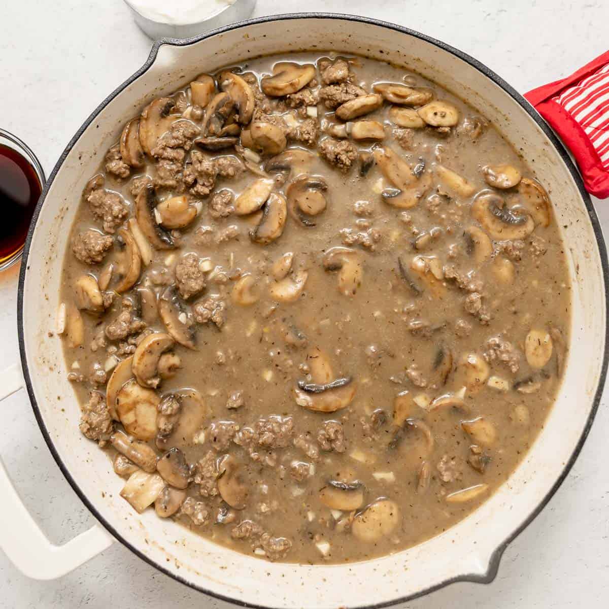 A white skillet filled with ground beef, sliced mushrooms, and brown gravy sits on a light countertop next to a red oven mitt.