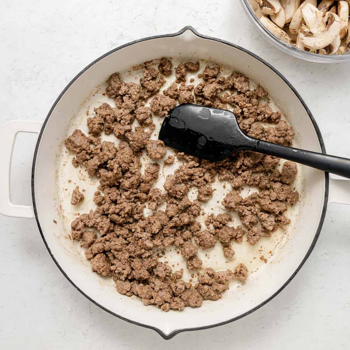 Ground beef is being browned in a white skillet with a black spatula; a bowl of sliced mushrooms is visible in the upper right corner.