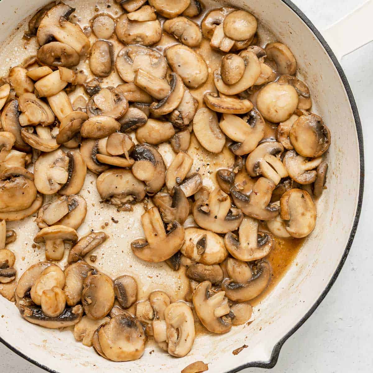 Sliced mushrooms sautéed in a white pan, showing light browning and some liquid at the bottom.