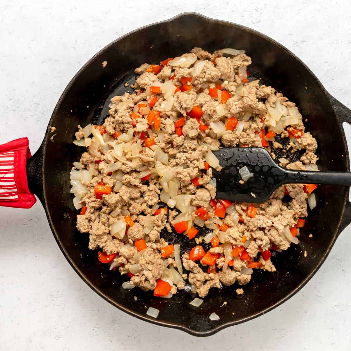Ground meat, chopped onions, and red bell peppers cooking in a black skillet, with a black spatula resting inside the pan.