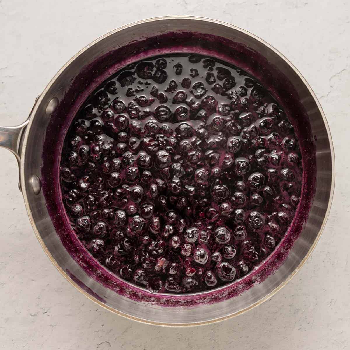 A stainless steel saucepan filled with simmering blueberry compote on a light gray surface.