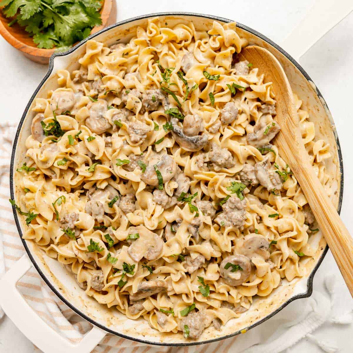 A skillet filled with creamy beef stroganoff pasta, garnished with chopped parsley, sits on a striped towel with a wooden spoon resting inside.