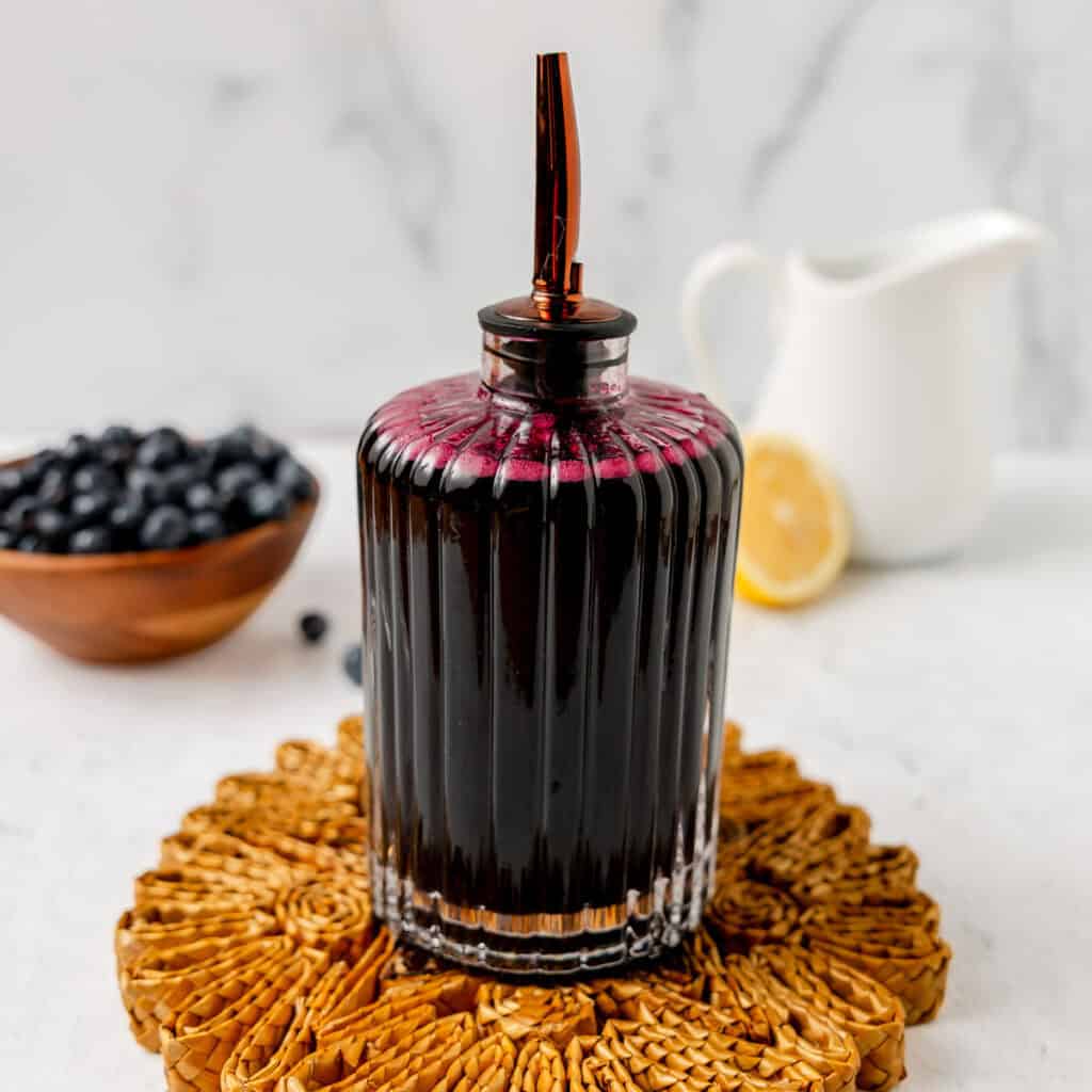 A glass bottle filled with dark blueberry syrup and a metal spout sits on a woven mat, with a bowl of blueberries, a lemon slice, and a white pitcher in the background.