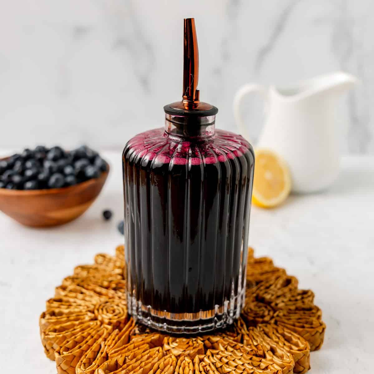 A glass bottle filled with dark blueberry syrup and a metal spout sits on a woven mat, with a bowl of blueberries, a lemon slice, and a white pitcher in the background.