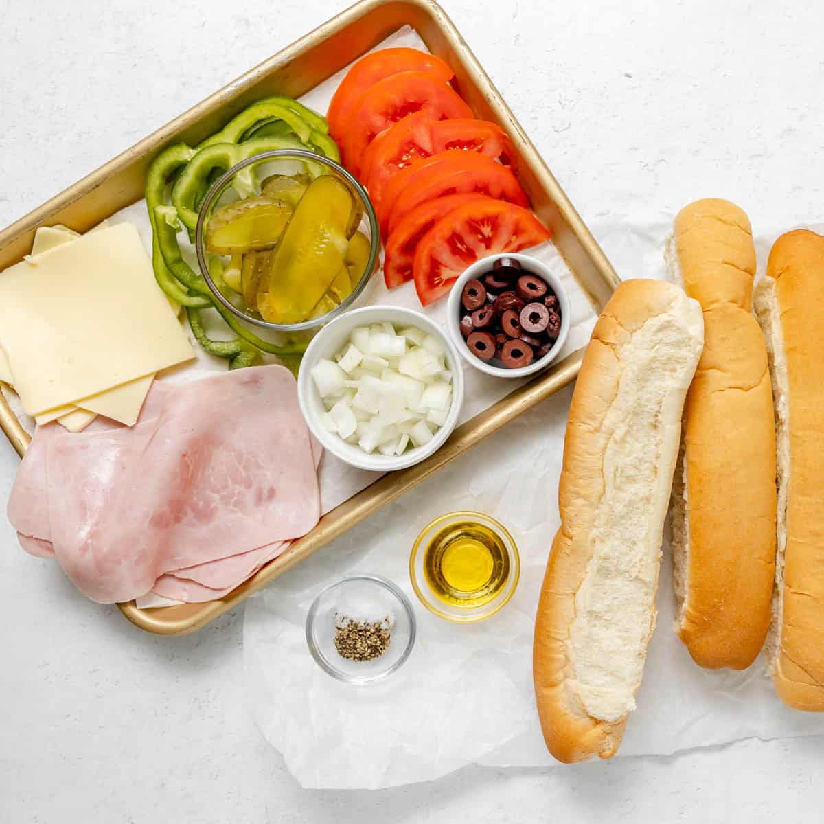 Tray with sliced ham, cheese, green peppers, tomatoes, pickles, onions, olives, spices, oil, and two hoagie rolls on a white surface.