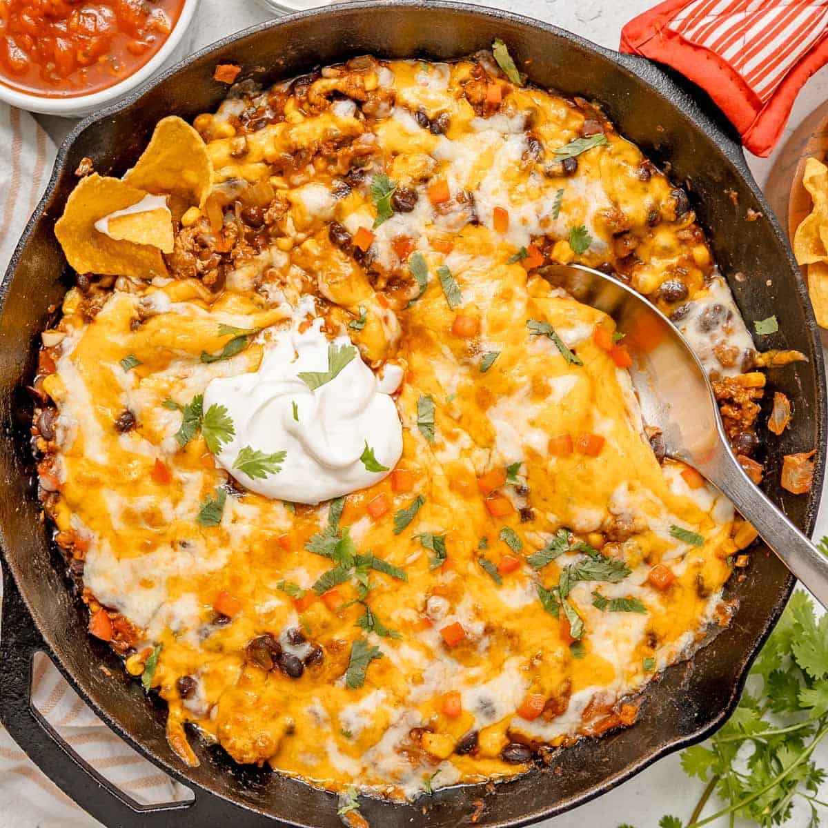 Skillet of cheesy taco casserole topped with sour cream, chopped tomatoes, and cilantro, with corn chips and a serving spoon visible.