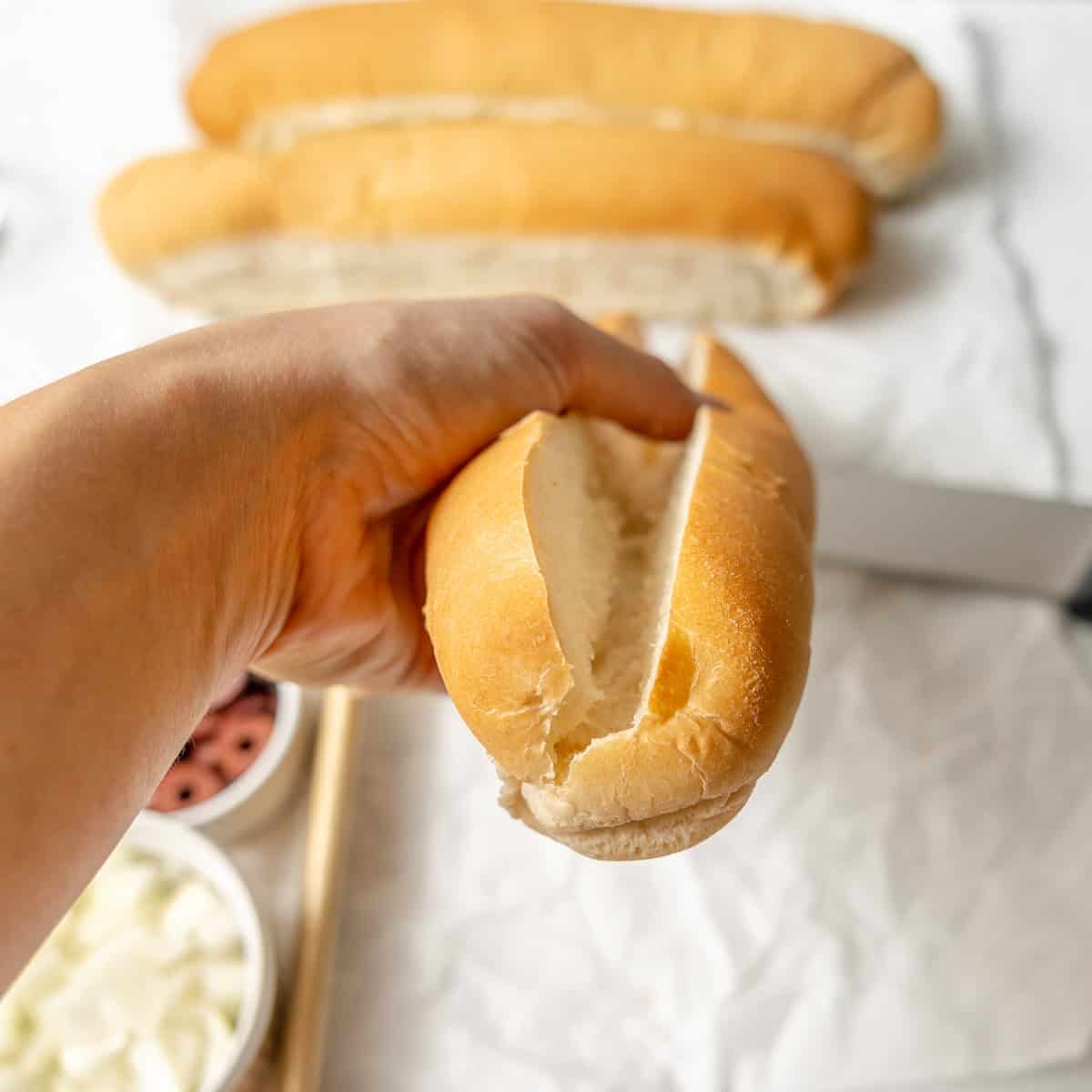 A hand holds a sliced sandwich roll above a surface with two more rolls in the background and small bowls of toppings nearby.