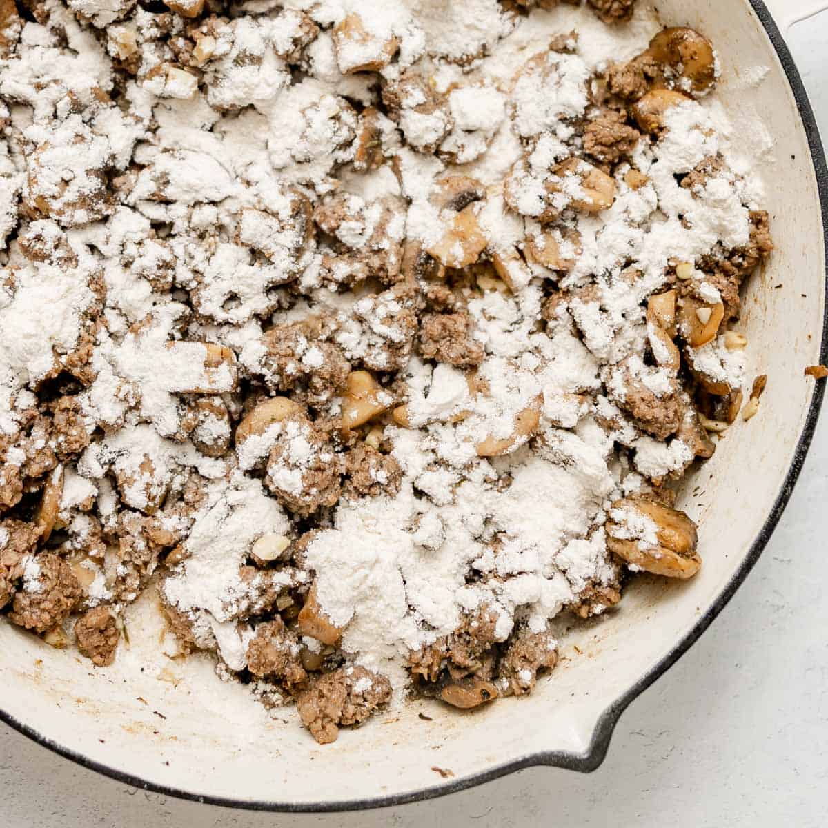 Ground beef and mushrooms cooking in a skillet, topped with a layer of white flour.