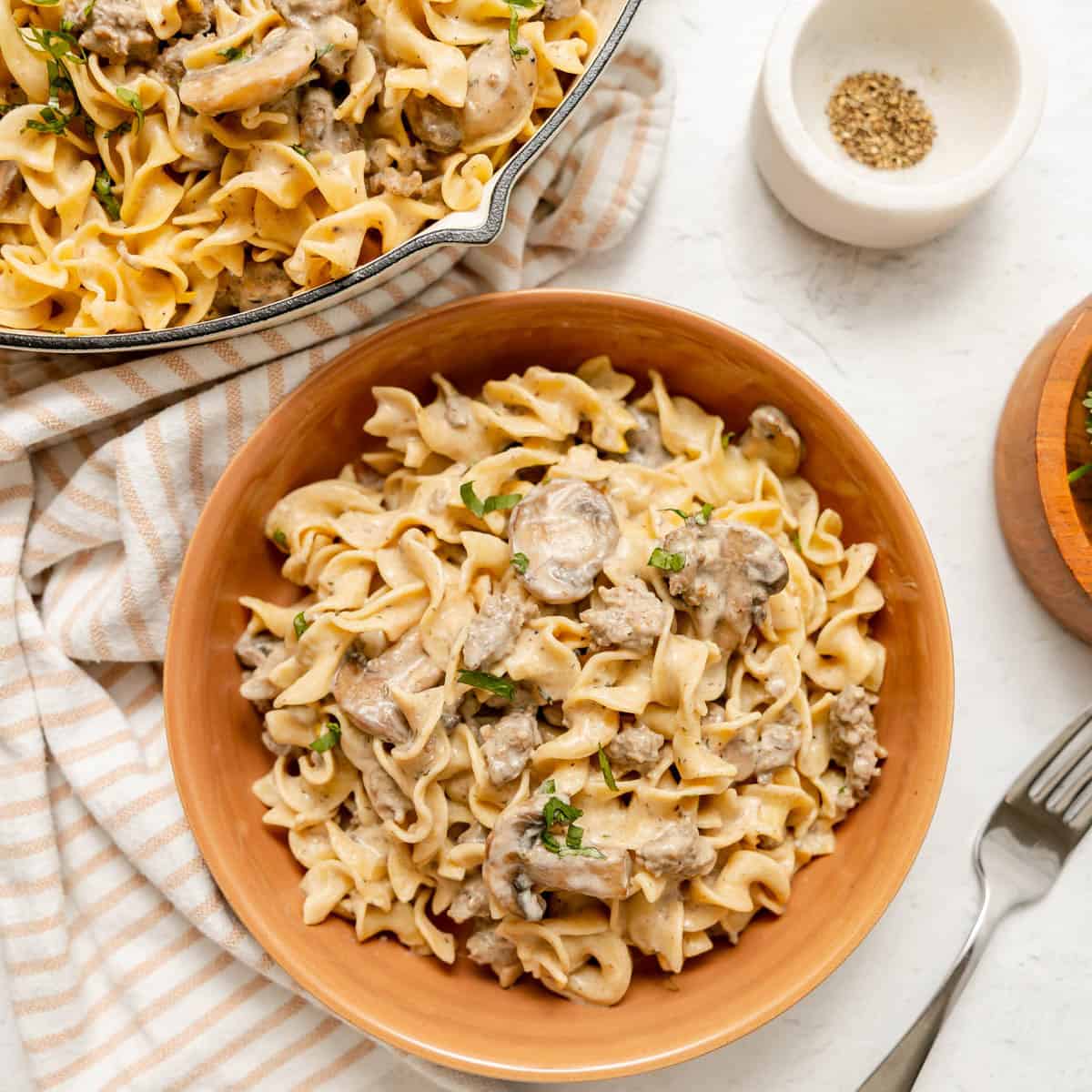 A bowl of creamy beef stroganoff with egg noodles and mushrooms, garnished with chopped herbs, next to a fork, napkin, and pepper in a small bowl.