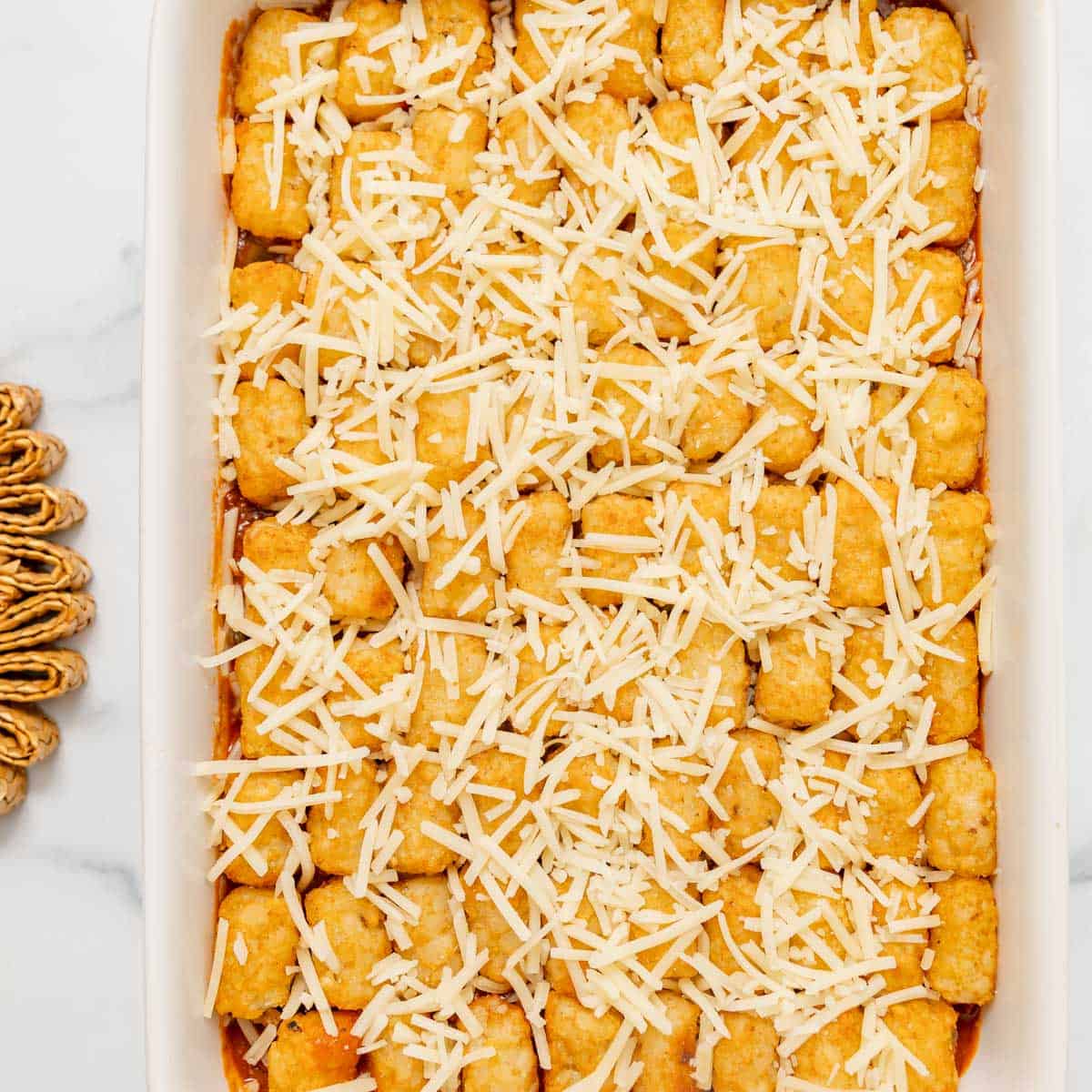 Rectangular casserole dish filled with tater tots arranged in rows and topped with shredded cheese, ready to be baked.