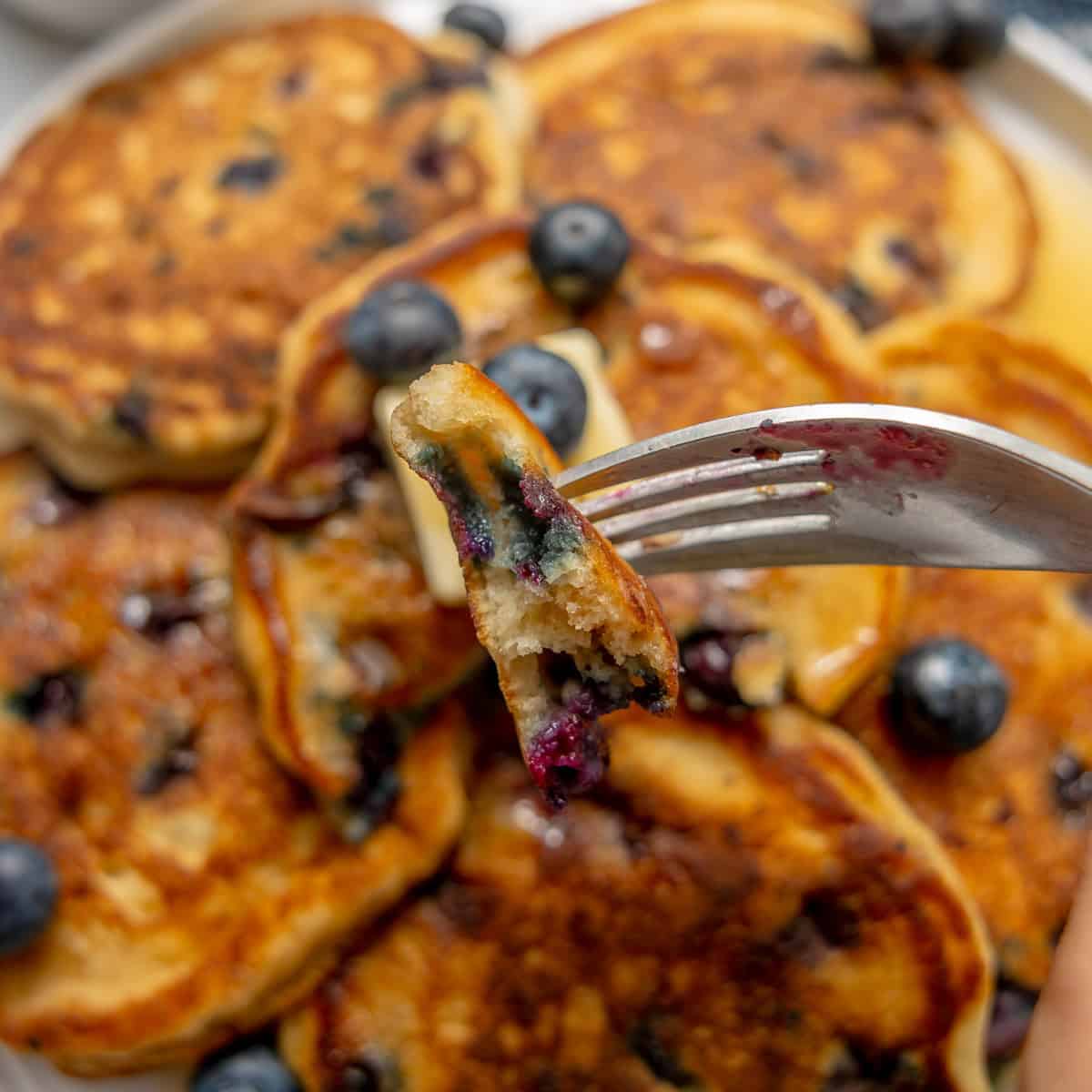 A fork holds a bite of blueberry pancake above a plate stacked with more blueberry pancakes and fresh blueberries.