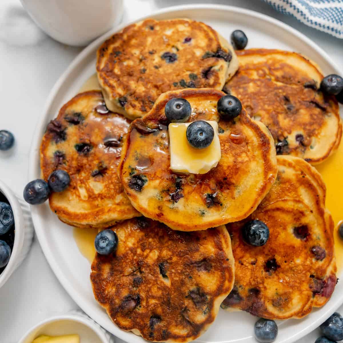 A plate of blueberry pancakes with butter, syrup, and fresh blueberries on top, surrounded by bowls of blueberries and maple syrup.