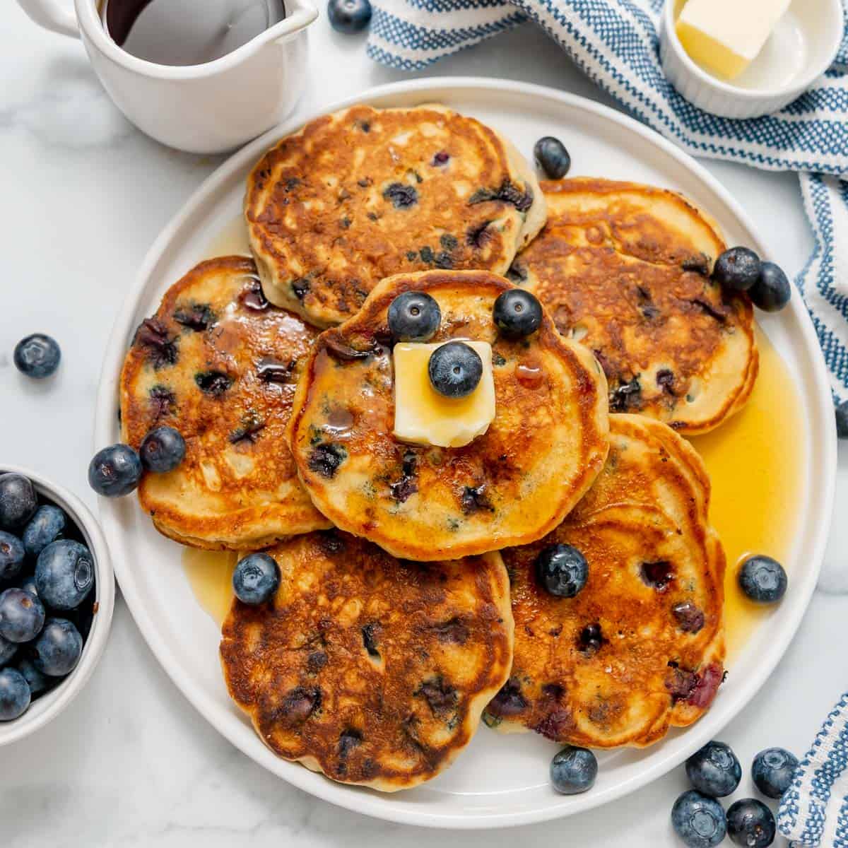 A plate of blueberry pancakes topped with butter and blueberries, surrounded by syrup, blueberries, and a cloth napkin on a white surface.