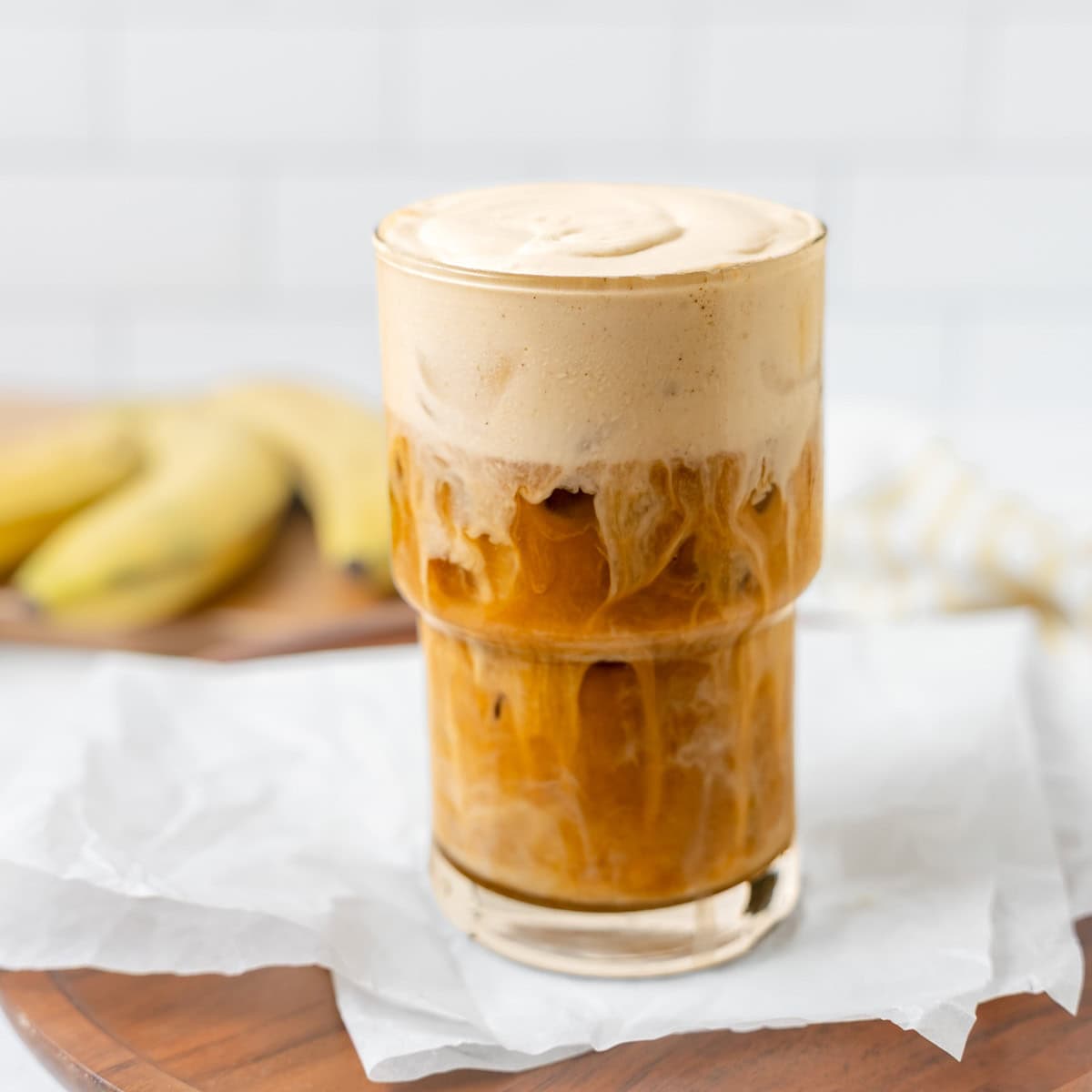 A glass of iced coffee topped with thick, creamy foam sits on parchment paper, with bananas and a striped cloth in the blurred background.
