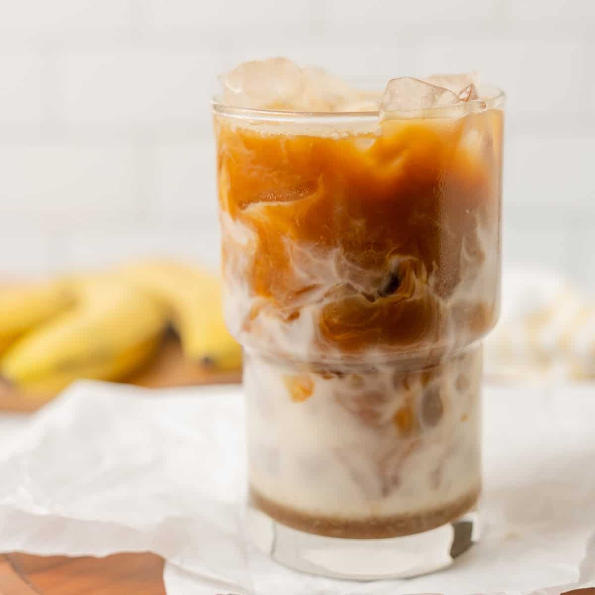A glass of iced coffee with milk swirling through it sits on a white surface, with bananas blurred in the background.