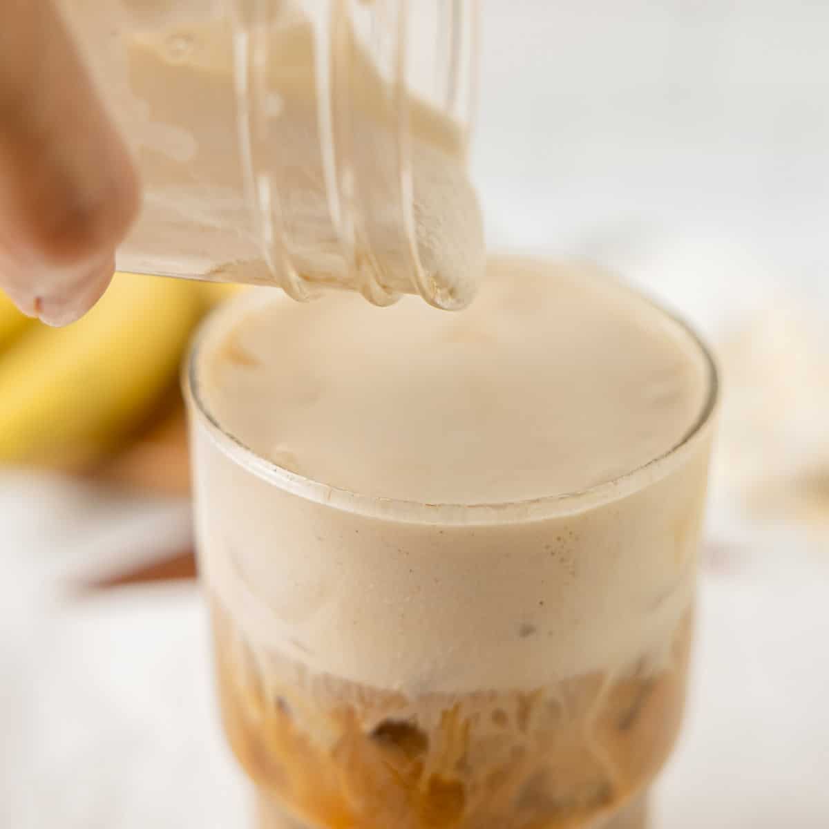 A hand pours frothy liquid from a jar into a glass filled with iced coffee on a light surface.