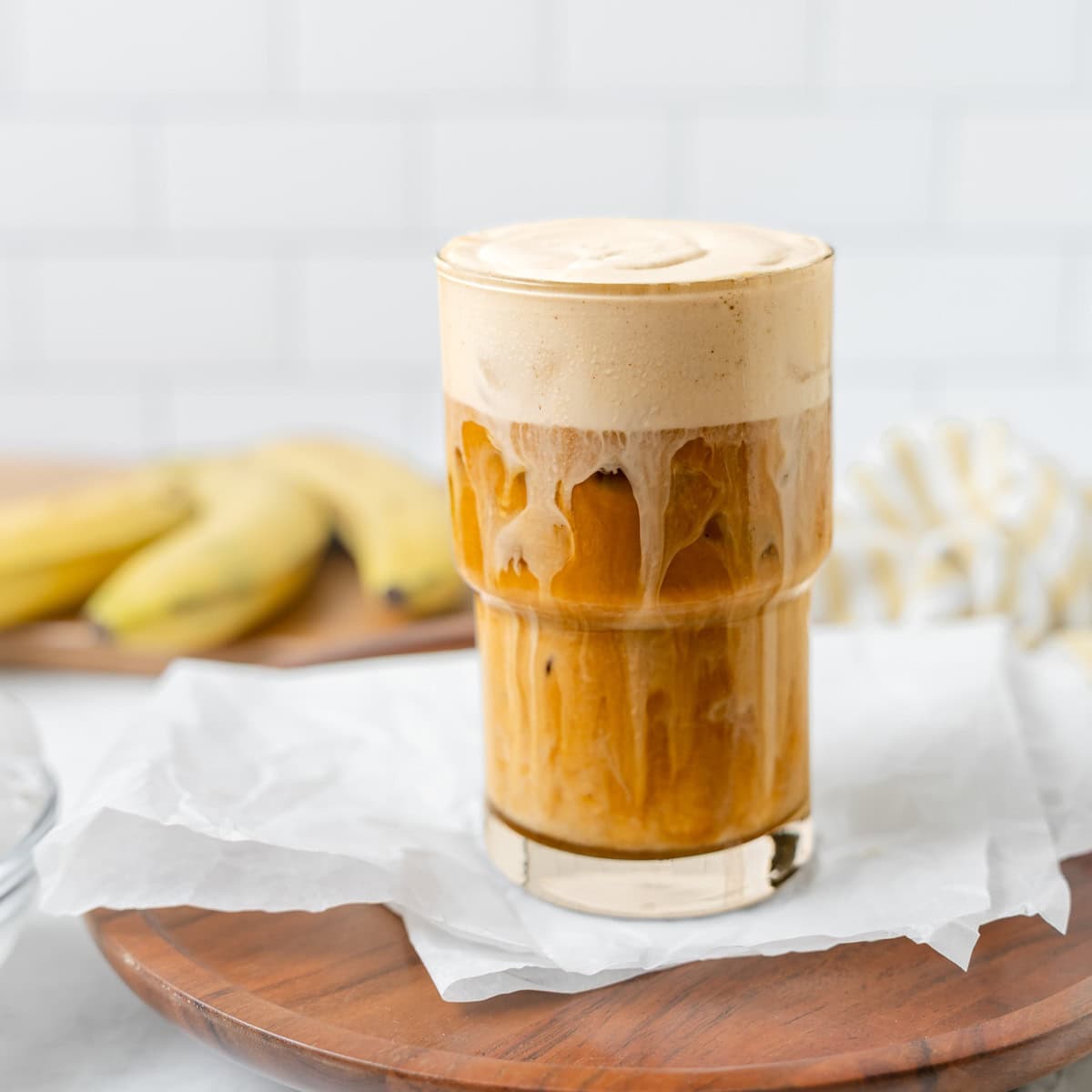 A glass of iced coffee with frothy foam on top sits on a piece of white parchment paper. Bananas are blurred in the background on a wooden board.