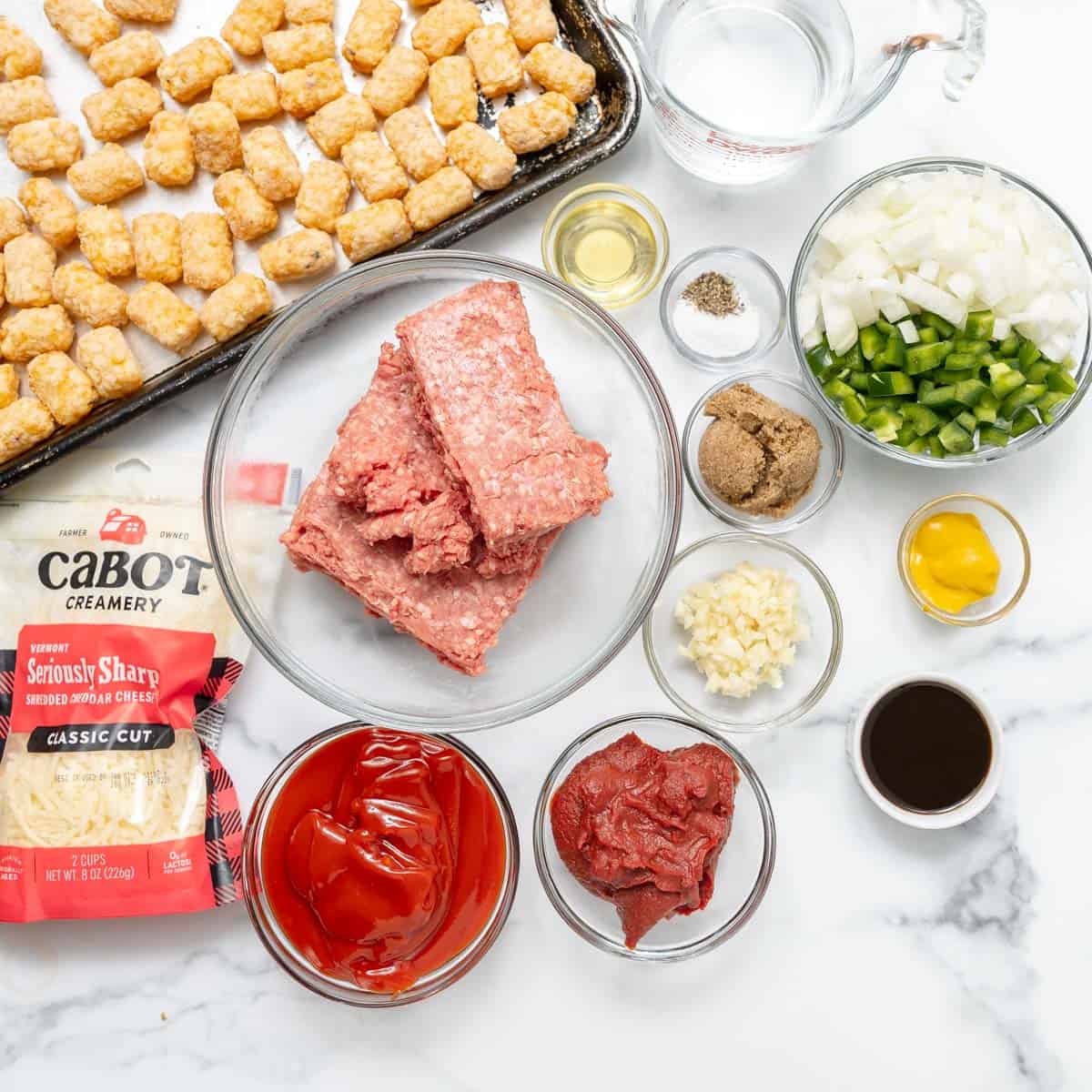 Ingredients for a tater tot casserole arranged on a white surface, including ground beef, vegetables, seasonings, cheese, ketchup, and tater tots on a baking tray.