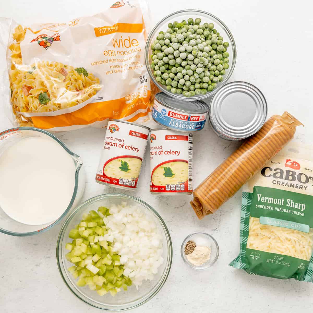 Ingredients for tuna casserole laid out on a white surface, including egg noodles, peas, canned tuna, cream of celery soup, milk, celery, onion, cheddar cheese, round crackers, and seasonings.