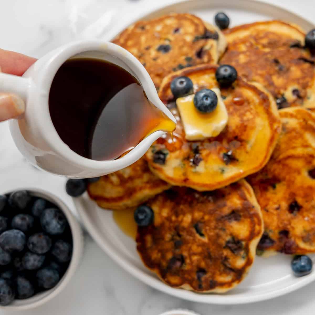 A hand pours syrup onto a stack of blueberry pancakes topped with butter and blueberries, with a bowl of blueberries nearby.