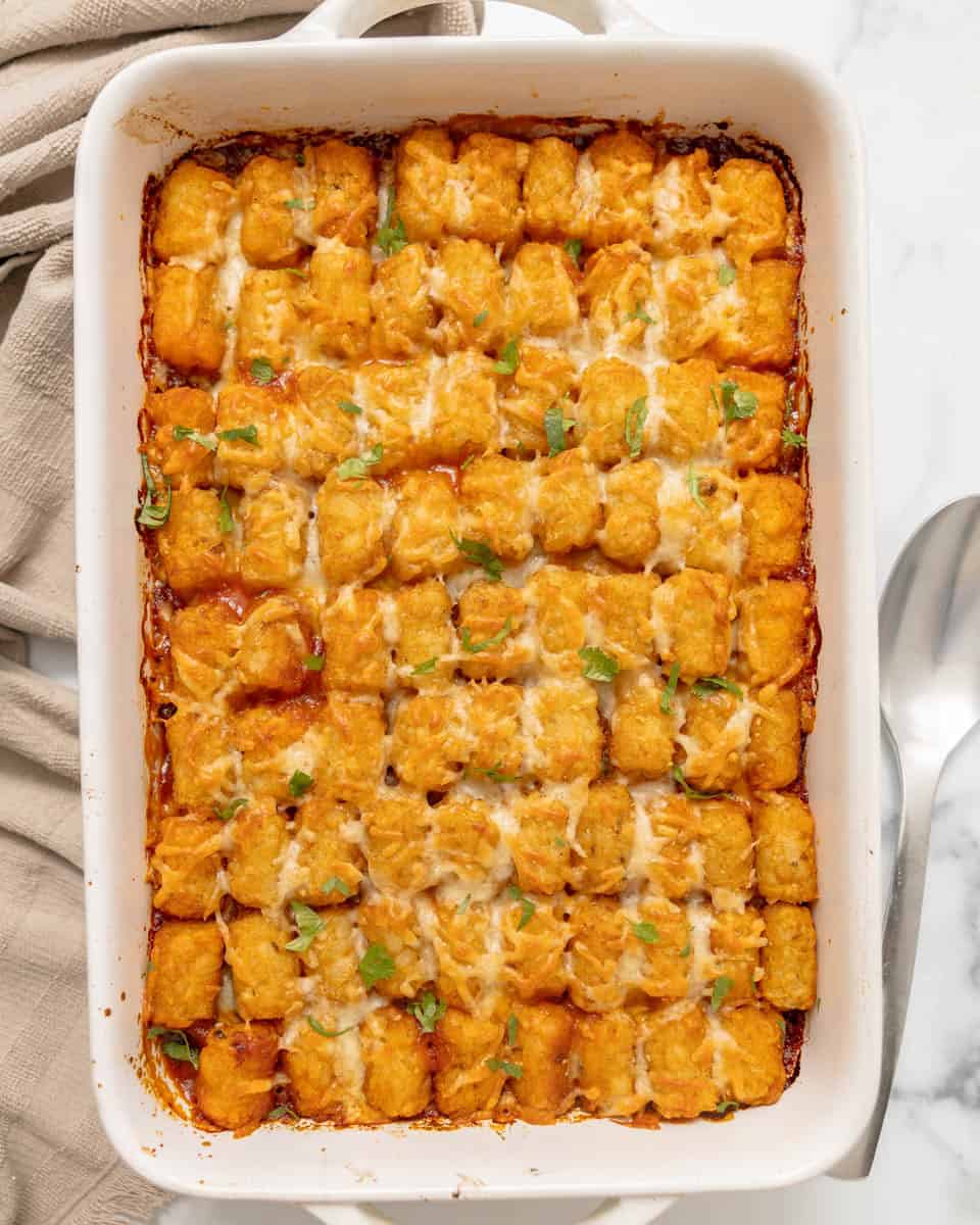 A baked casserole topped with rows of golden tater tots, melted cheese, and garnished with green herbs in a white dish.
