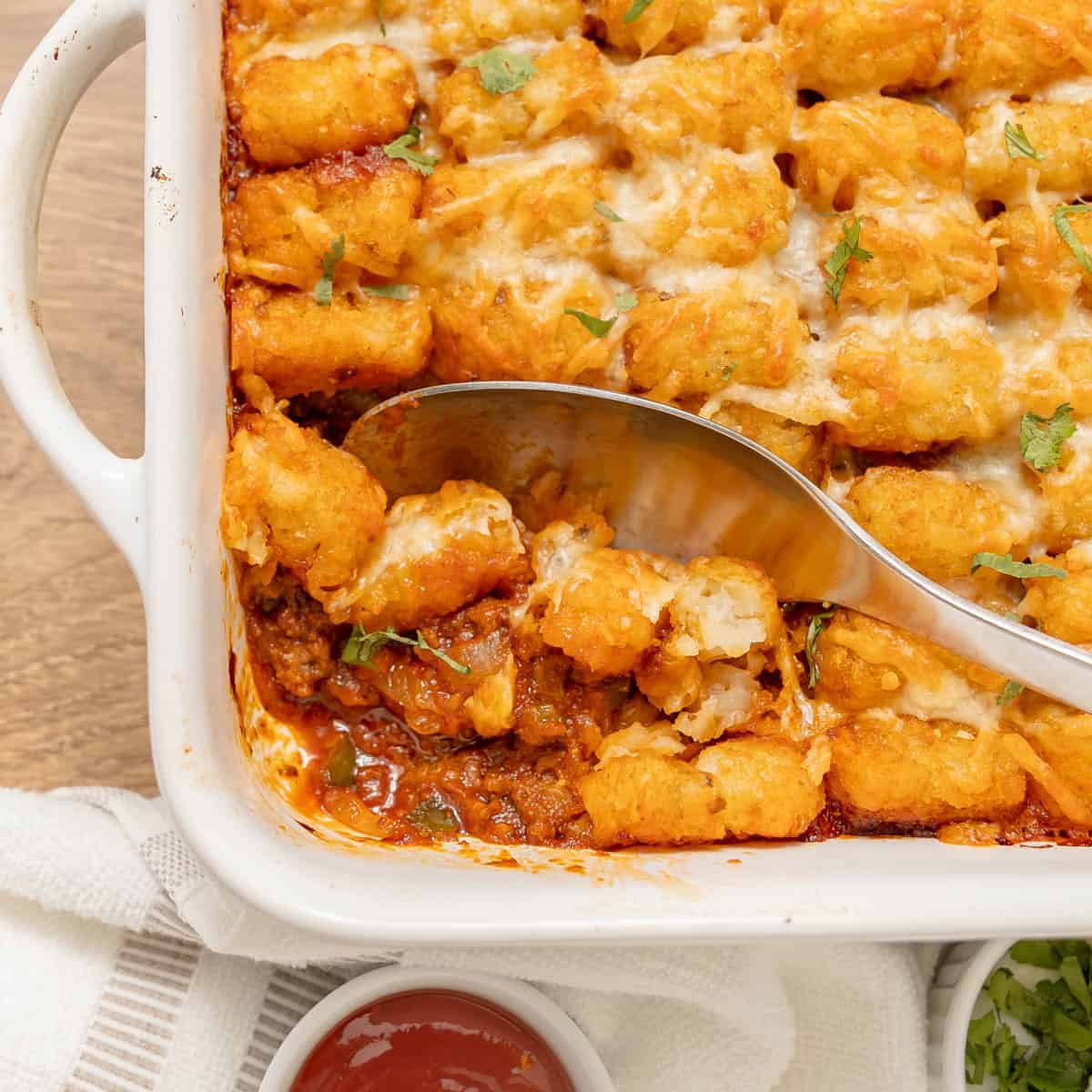 A baked tater tot casserole in a white dish with a metal spoon scooping a portion, showing ground meat and melted cheese.