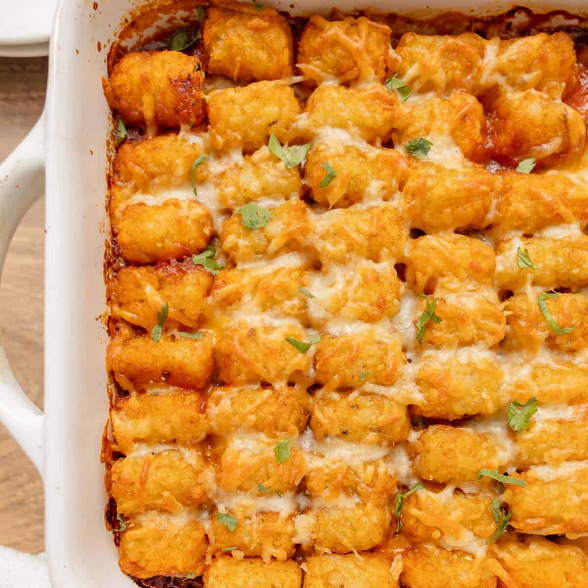 A baked tater tot casserole in a white dish, topped with melted cheese and sprinkled with chopped herbs.
