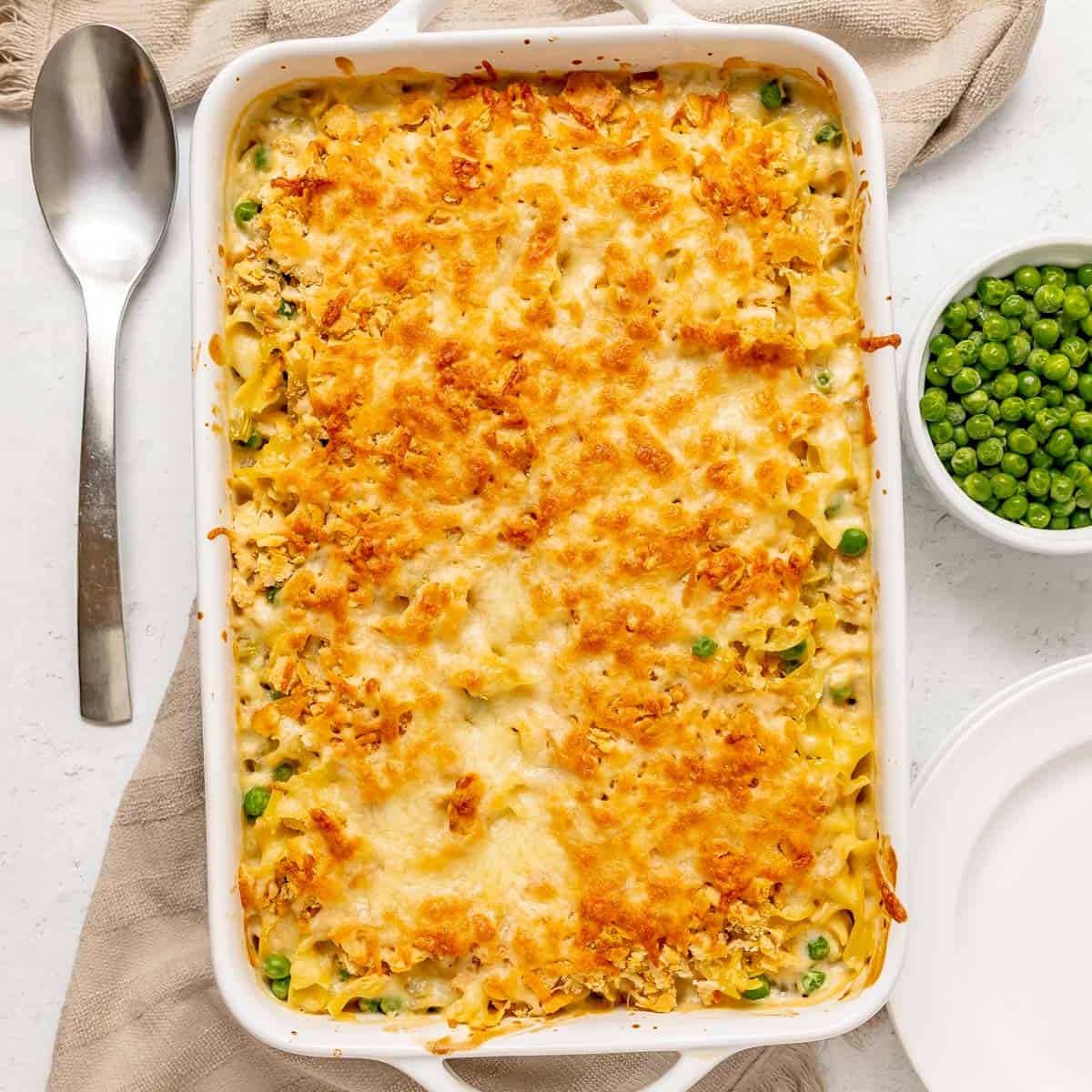 A baked casserole topped with melted cheese sits next to a spoon, a bowl of green peas, and an empty plate on a white surface.