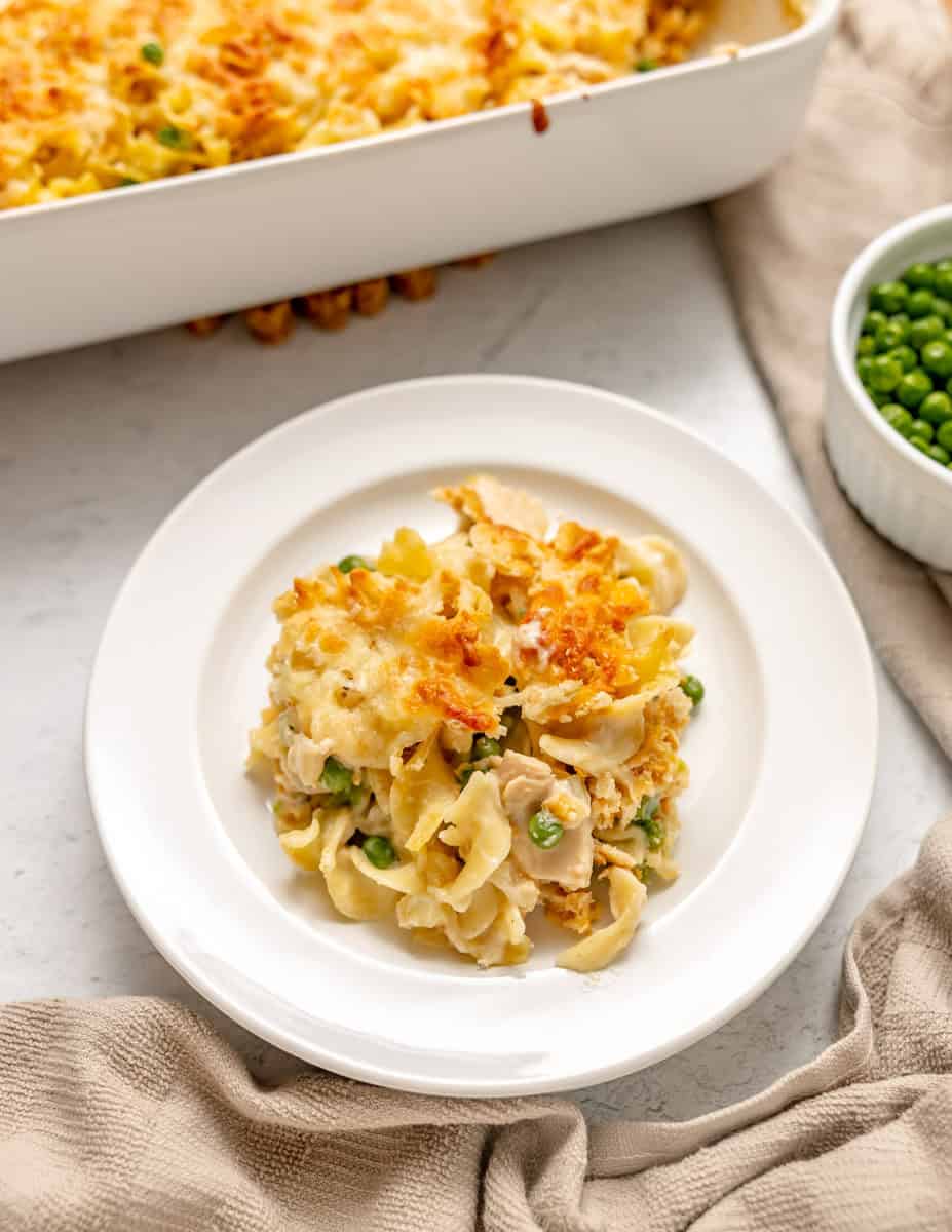 A serving of baked casserole with noodles, chicken, peas, and melted cheese on a white plate, next to a dish of green peas and a casserole dish.
