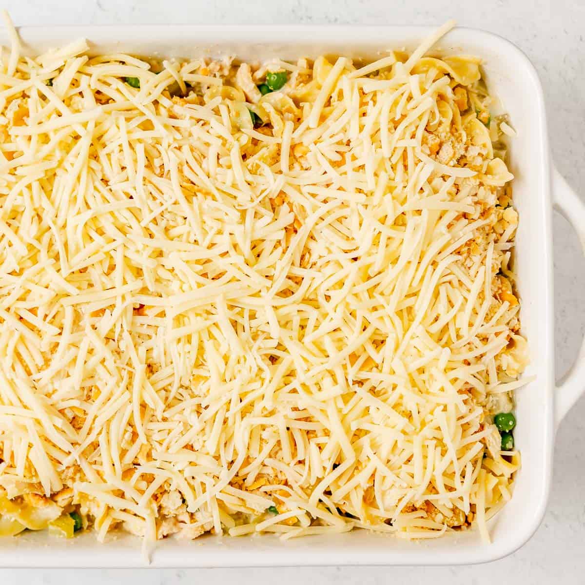 Rectangular white baking dish filled with an uncooked casserole topped with shredded cheese and breadcrumbs, ready to be baked.