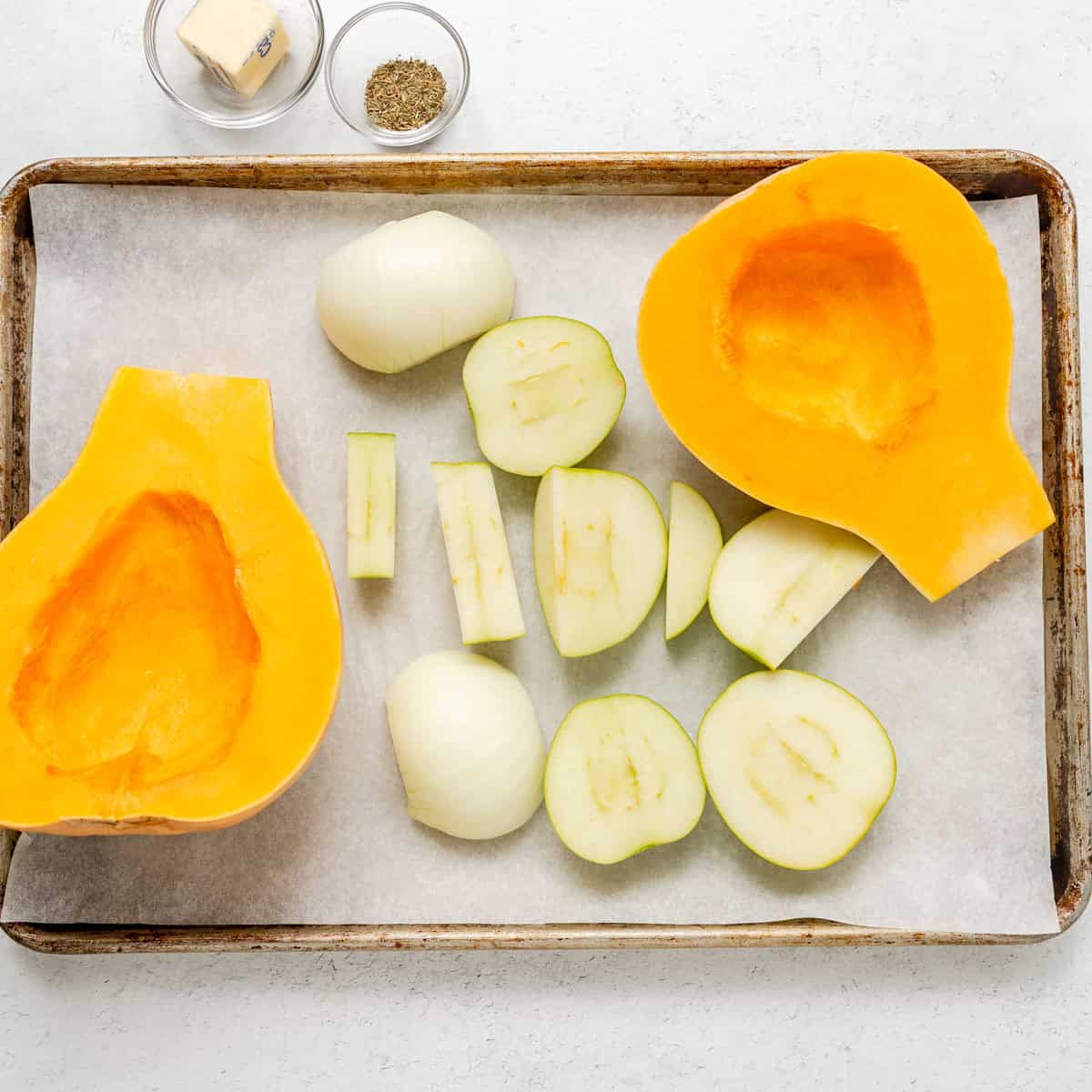 Sliced butternut squash, sliced apples, and onions on a parchment-lined baking sheet, with bowls of butter and black pepper nearby.
