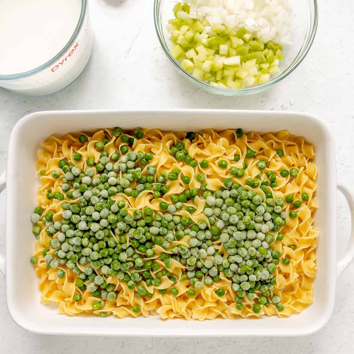 A white baking dish with egg noodles and frozen peas, next to bowls of milk, chopped onion, and chopped celery on a light countertop.